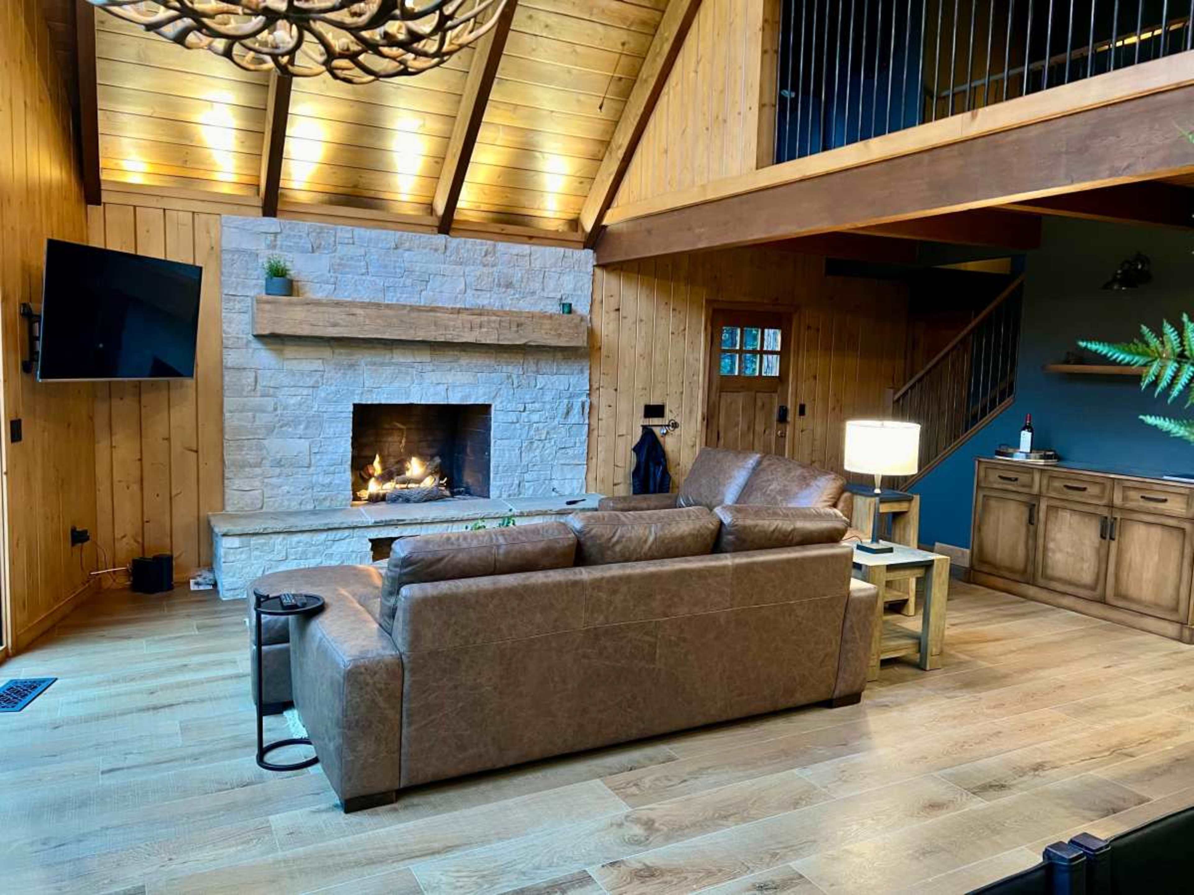The interior of a cozy cabin features a leather sofa facing a stone fireplace beneath a wooden beam ceiling.