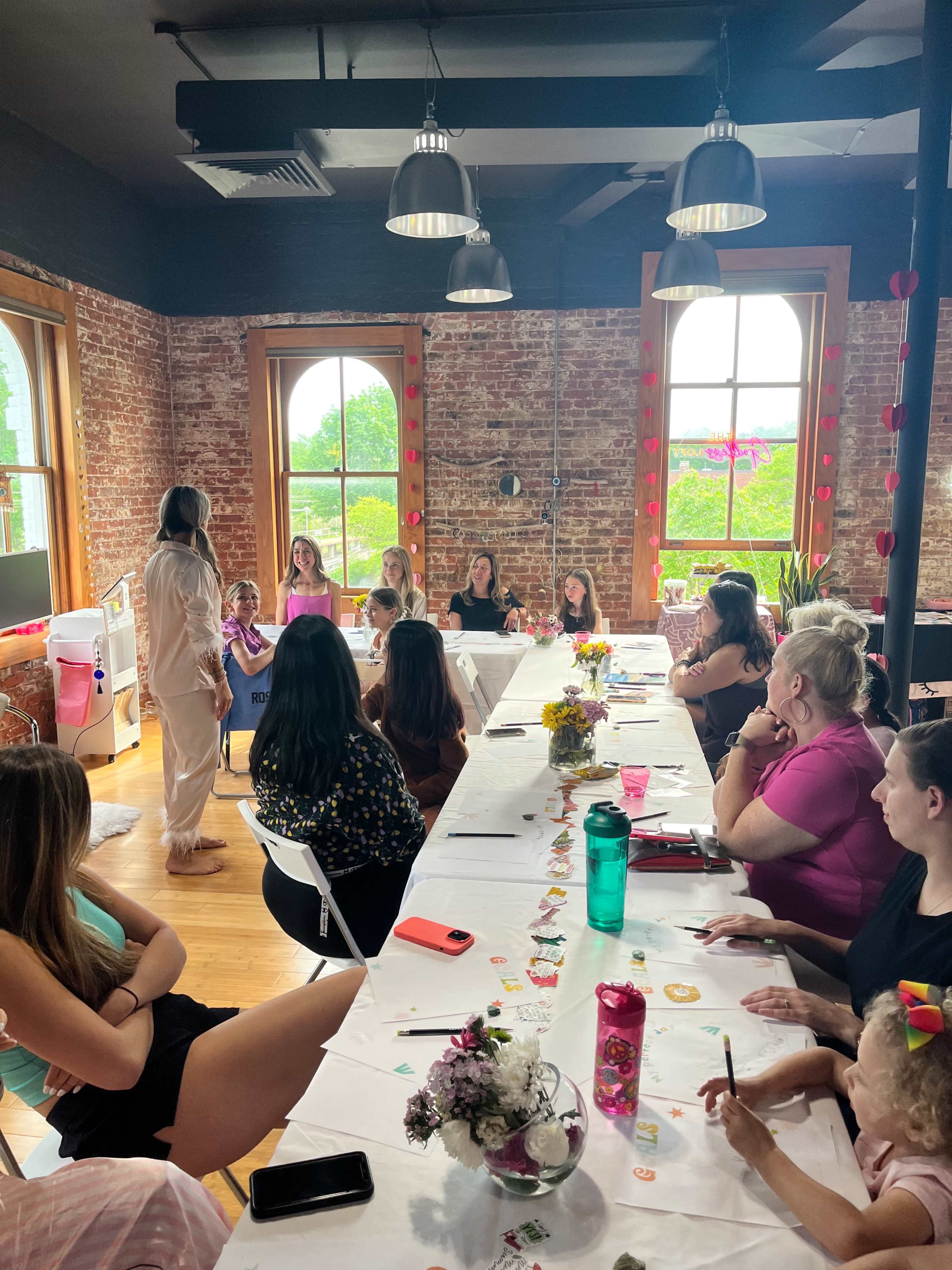 A group of women and children gather around a long table in a room with brick walls and large windows, engaging in a discussion or activity.