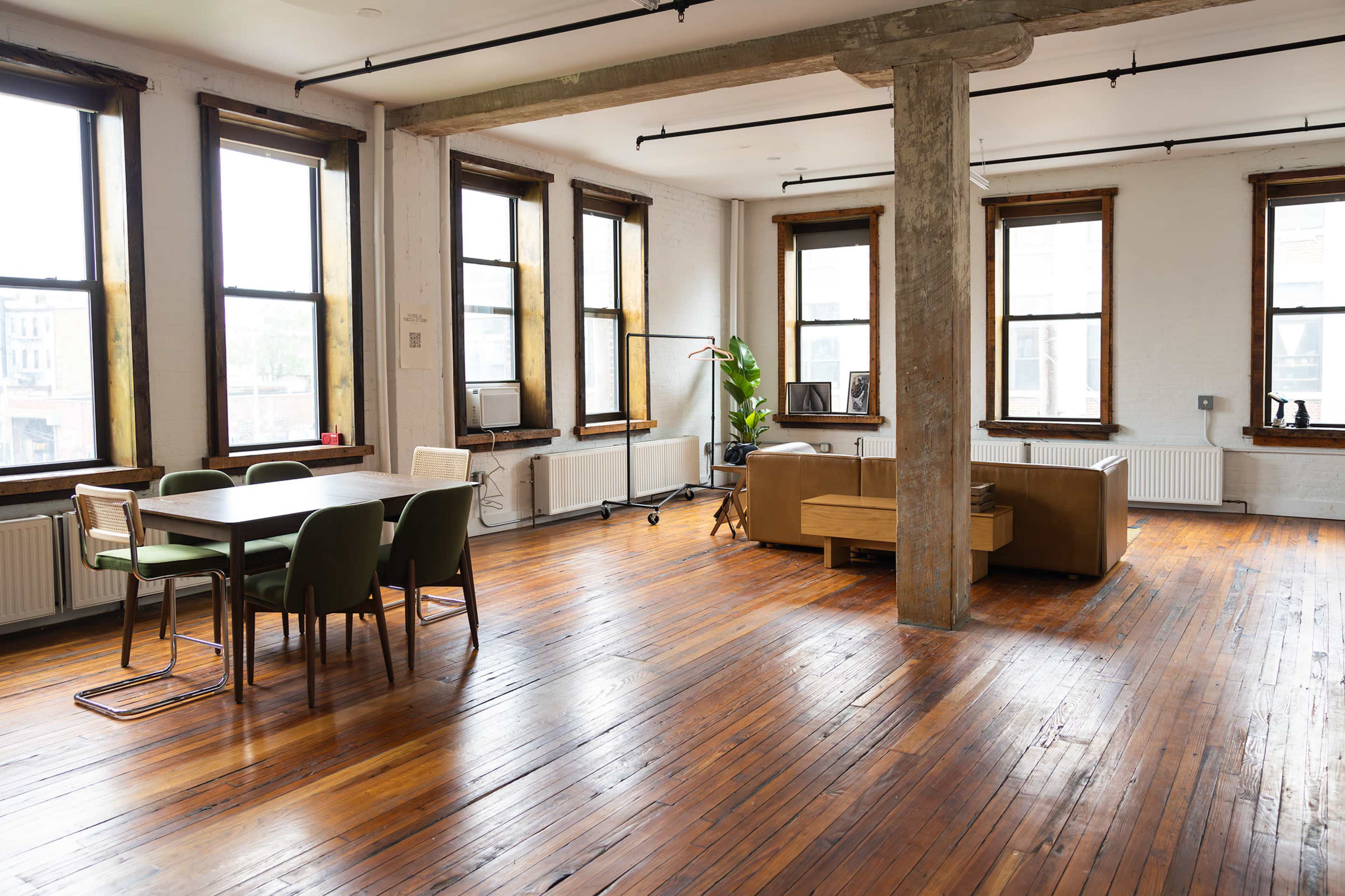 A spacious room with large windows, hardwood floors, a dining table with chairs, and a seating area featuring a sofa and a plant.