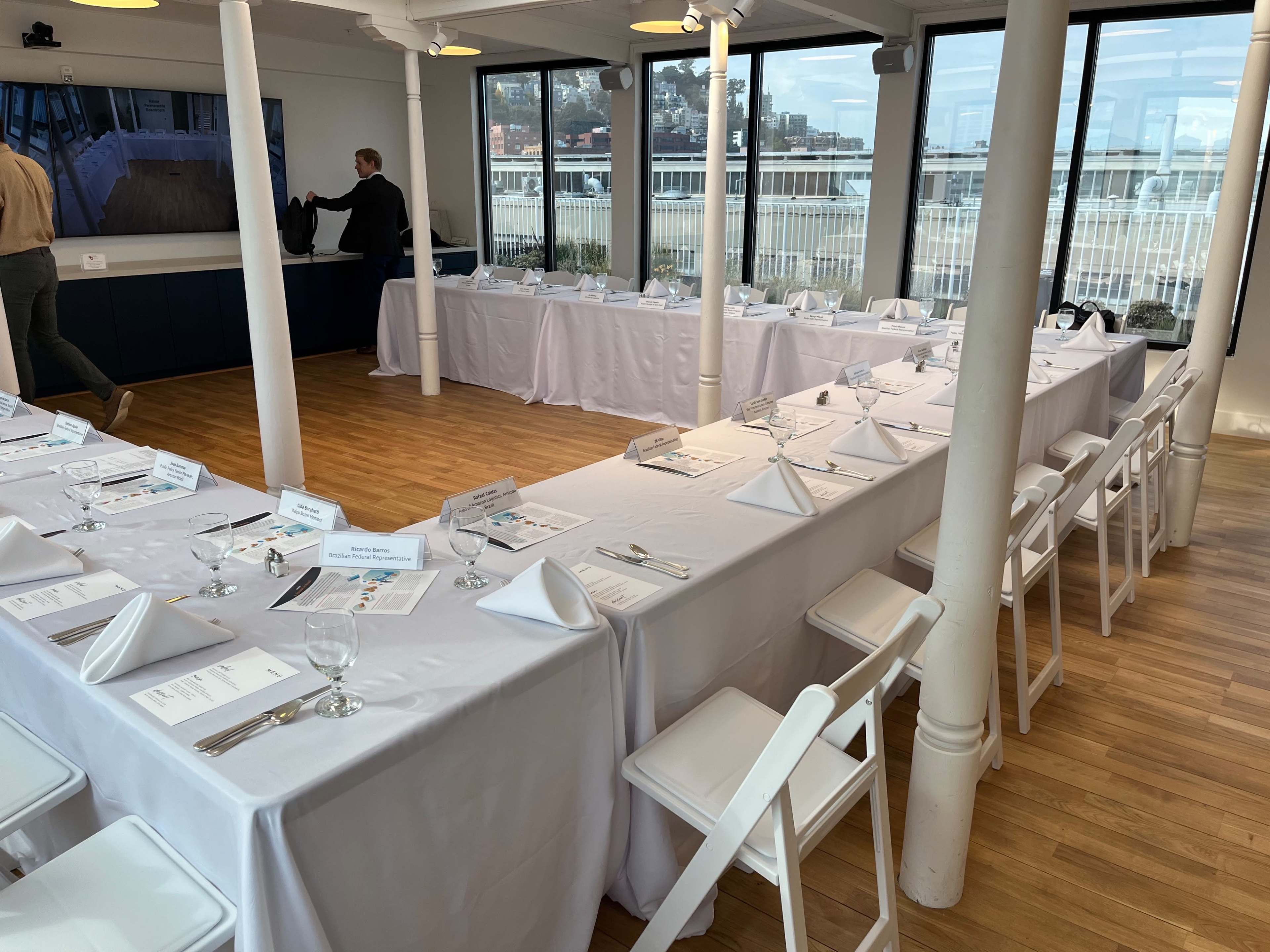 A spacious dining area features multiple tables set with white tablecloths and place settings, overlooking a waterfront view through large windows.