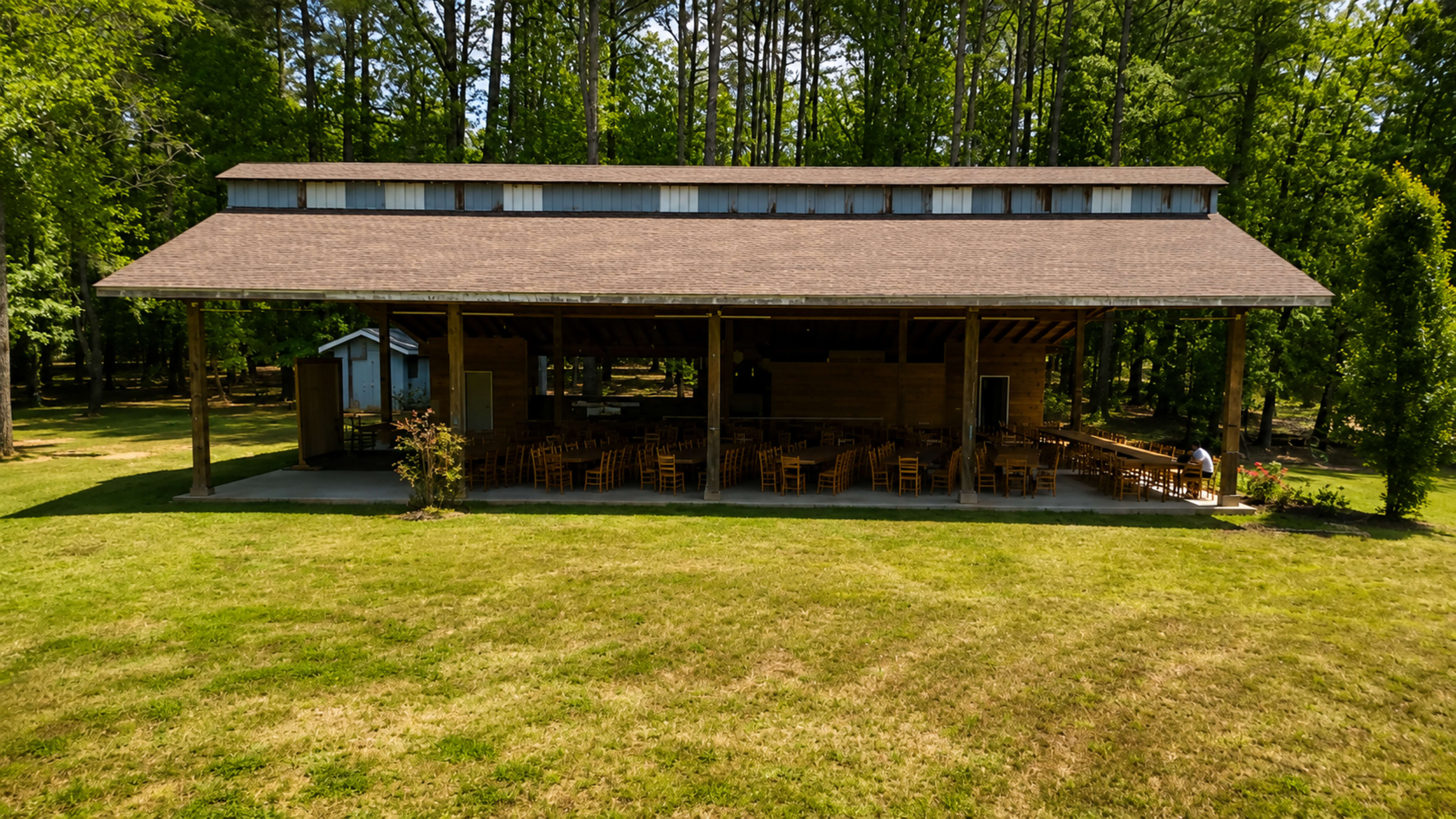 A large open pavilion with a sloped roof is surrounded by trees and has rows of wooden chairs set up inside.