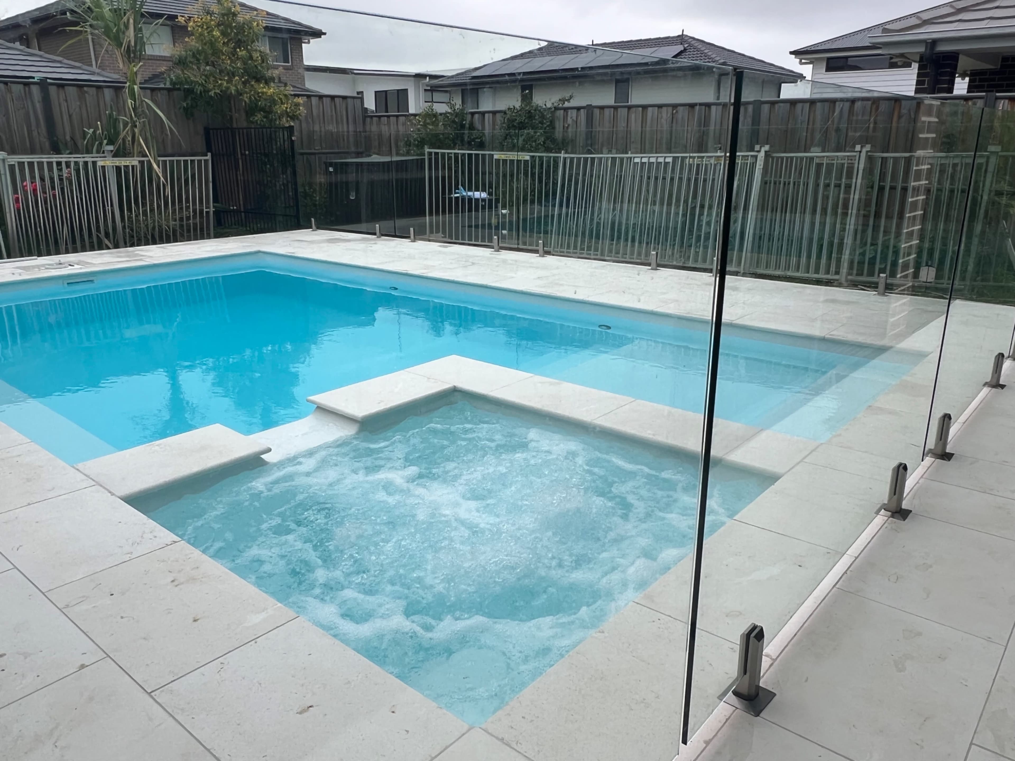 The image shows a residential swimming pool area with a clear blue pool featuring a spa section, surrounded by a glass fence.