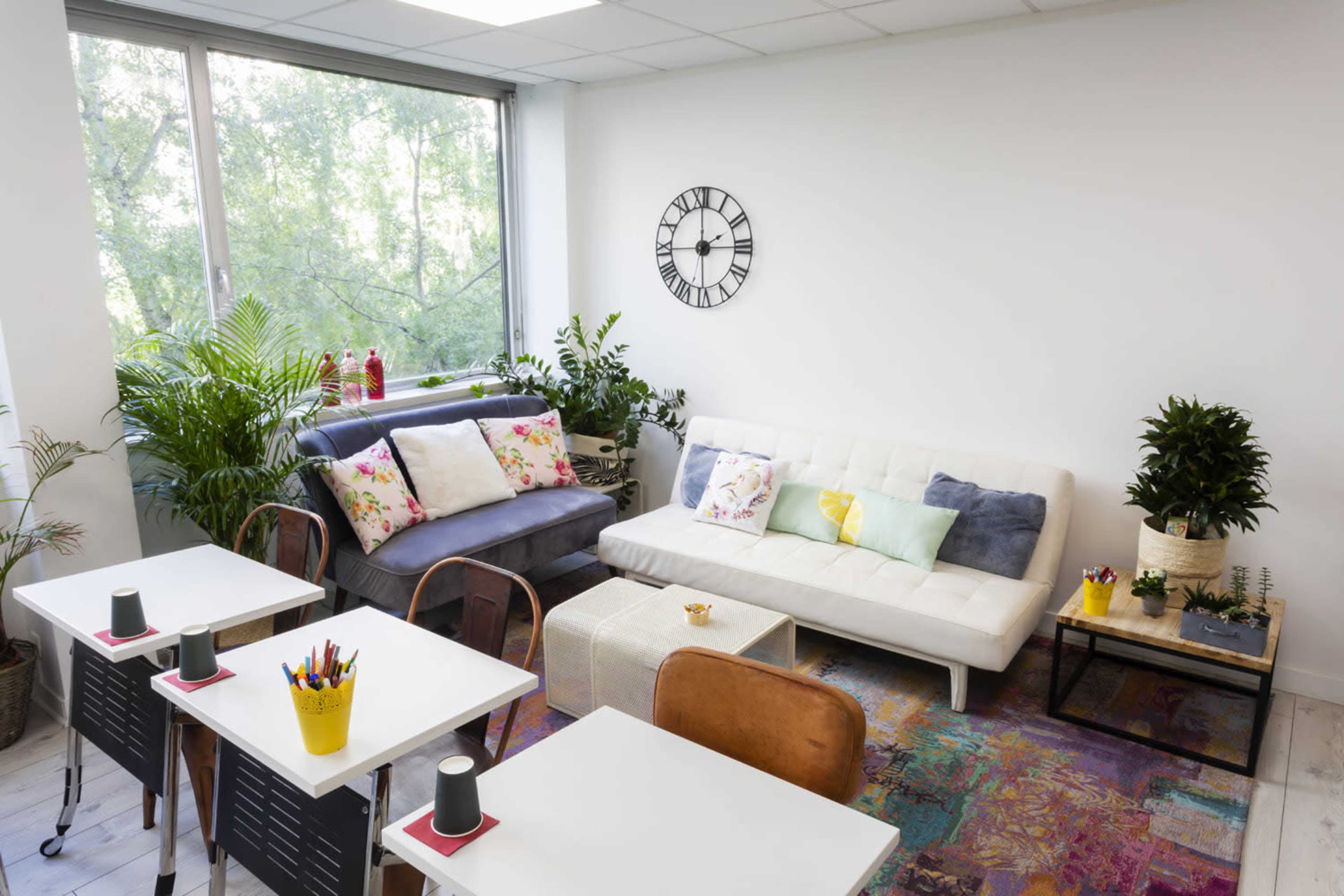A bright, modern lounge area with two sofas, a coffee table, potted plants, and a wall clock.