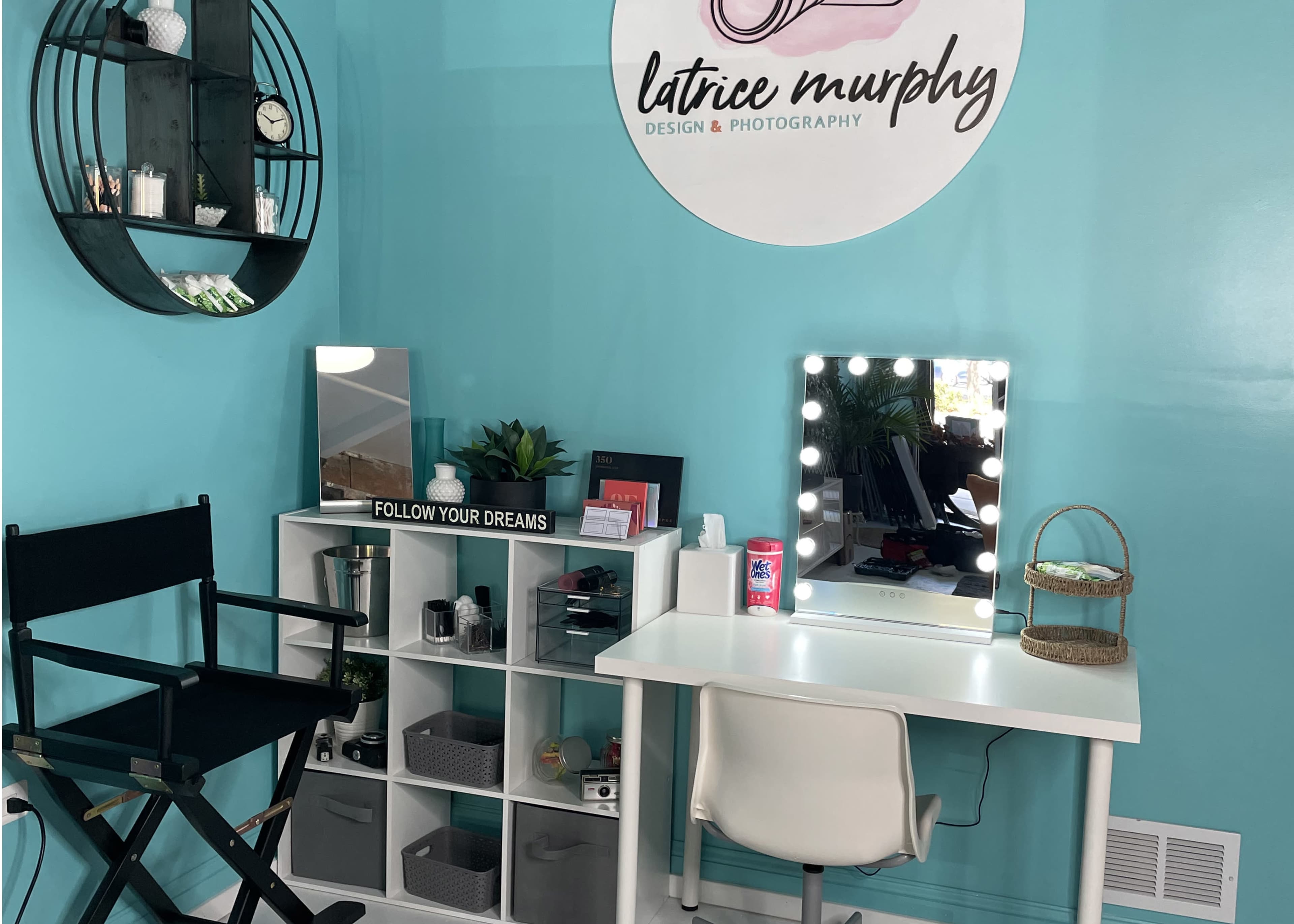 The image shows a teal wall with a round sign displaying "latrice murphy DESIGN & PHOTOGRAPHY," a makeup vanity with lights, a director's chair, a small shelf with organized items, and a motivational sign that says "FOLLOW YOUR DREAMS."