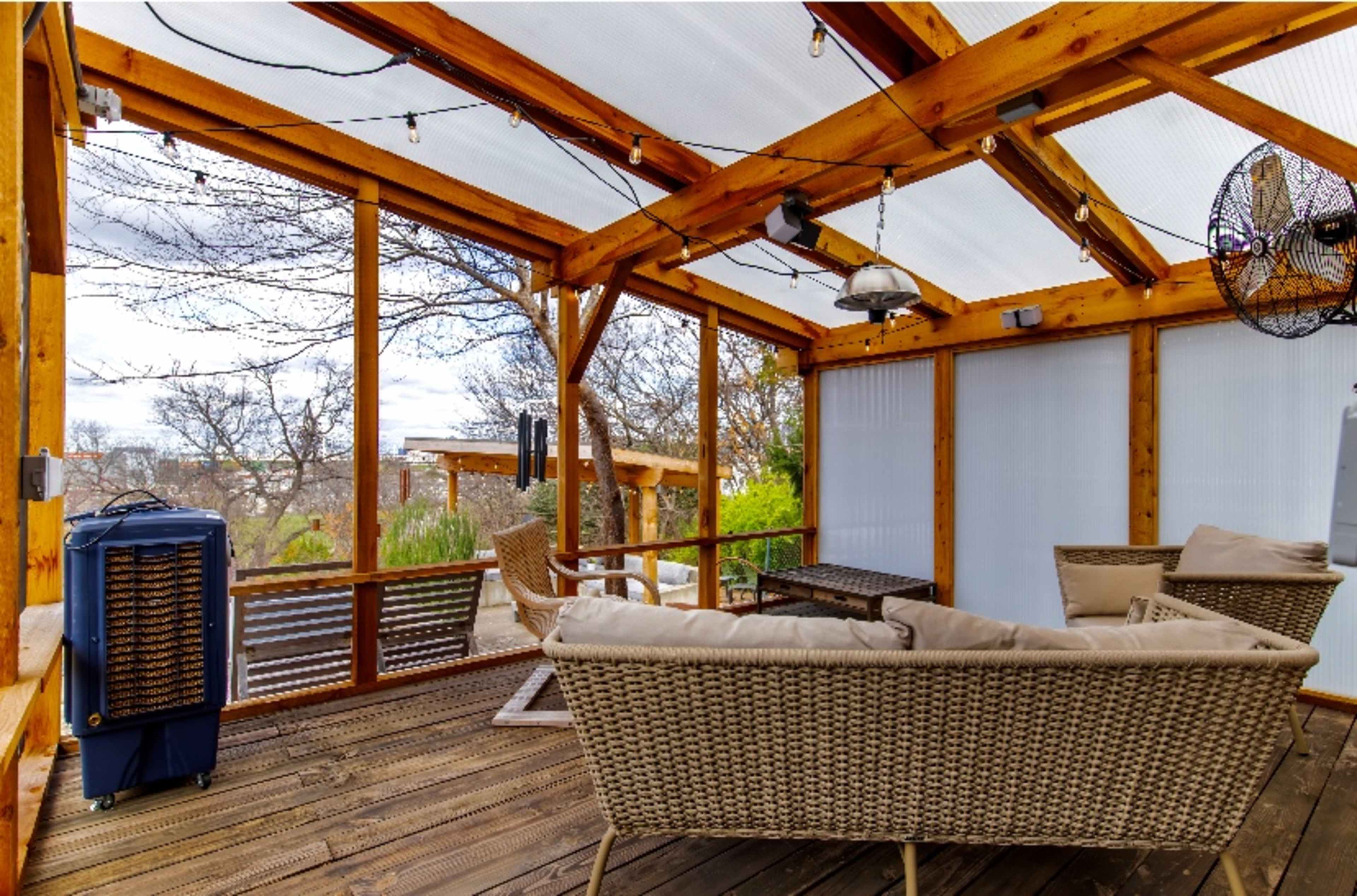 The image shows a wooden decked patio with a glass roof, featuring a seating area with wicker furniture and a portable heater.