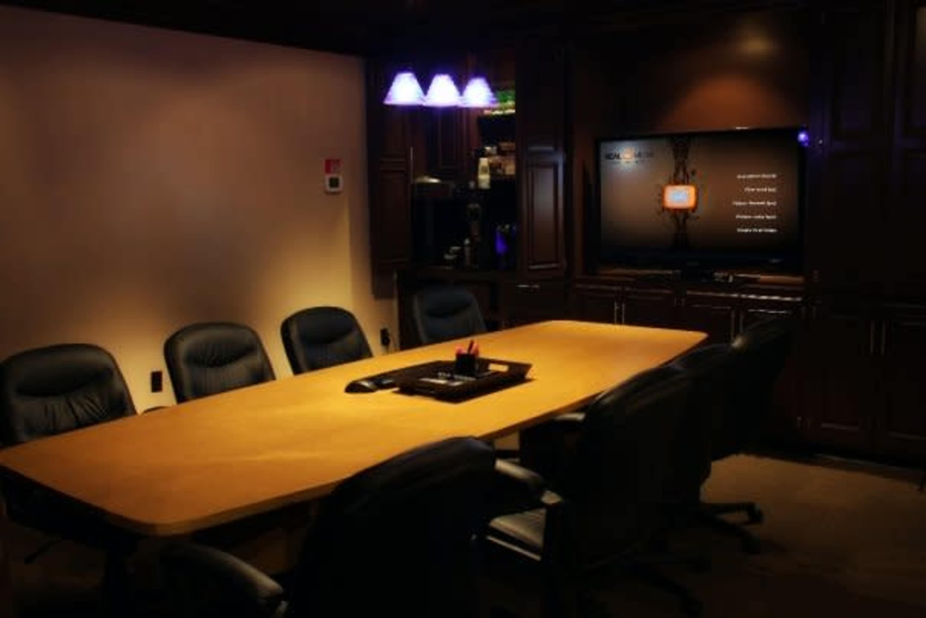 A large wooden conference table surrounded by black chairs sits in a dimly lit boardroom with a television screen displaying a menu on the wall.
