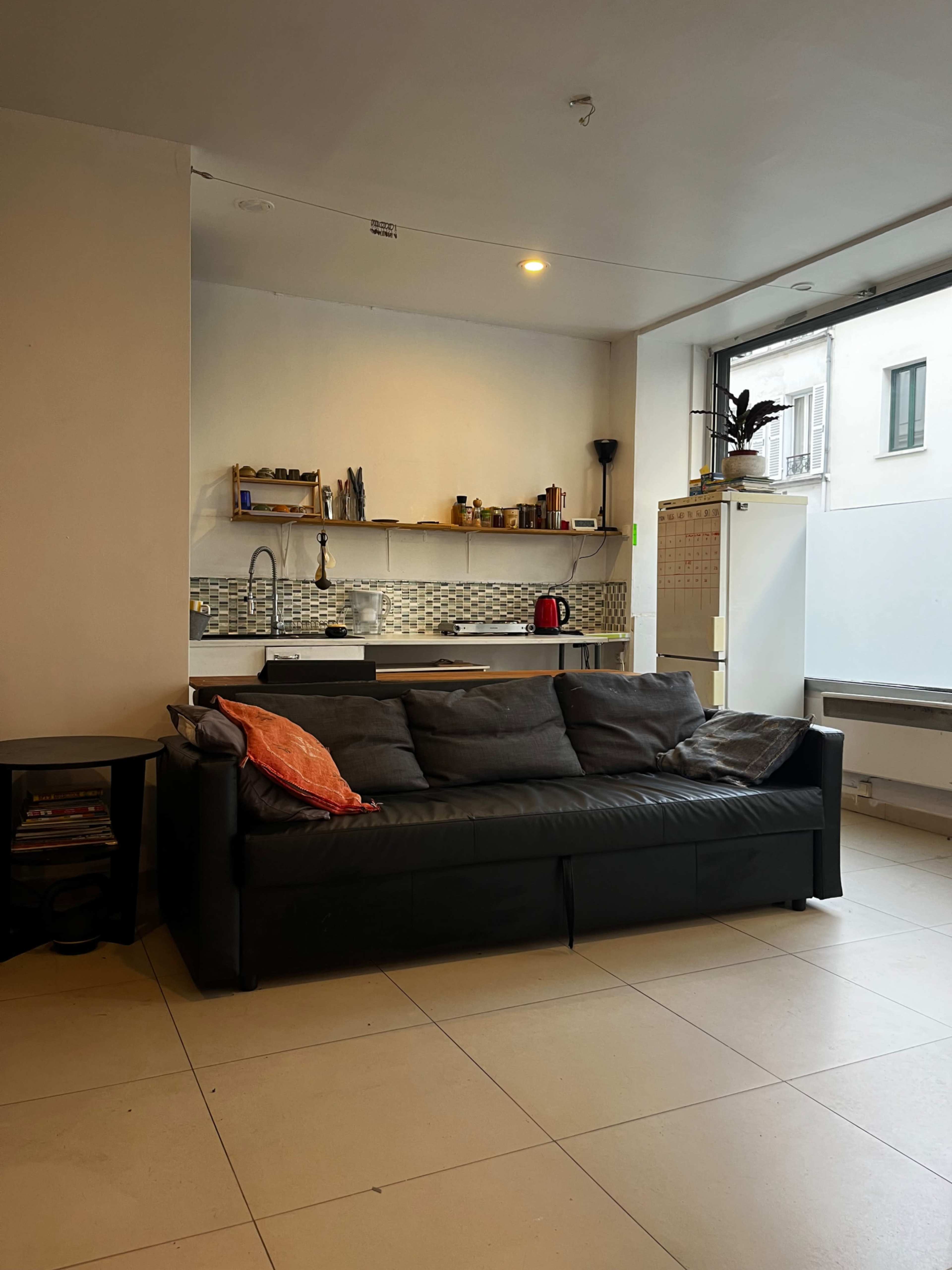 The image shows a modern living space featuring a black sofa with an orange pillow, a small kitchen area with shelves, a sink, and a refrigerator.
