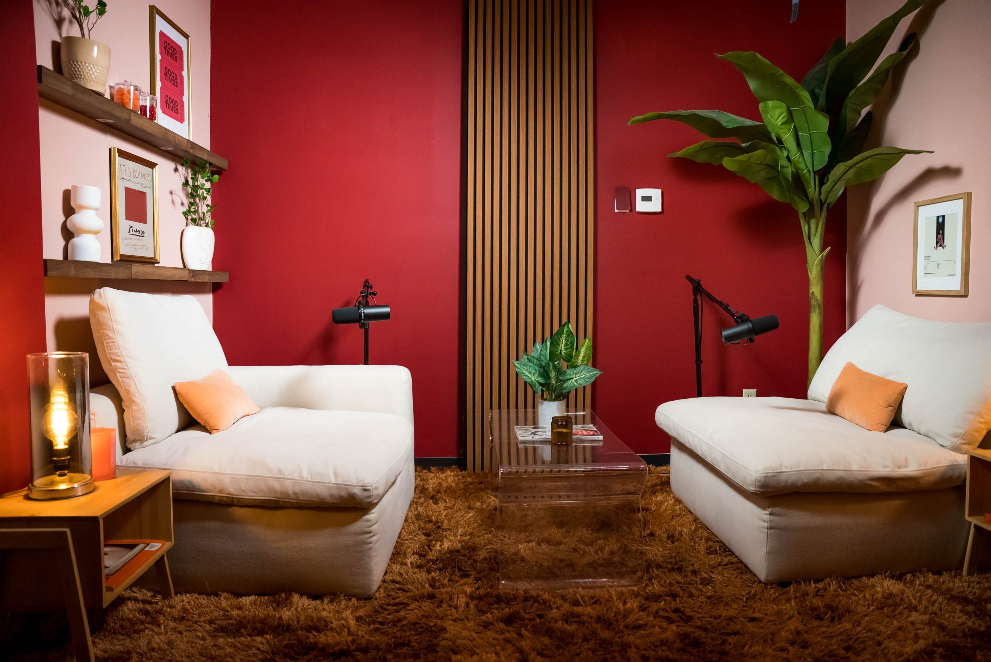 A cozy living room setup featuring two white armchairs, a small glass coffee table, a large potted plant, and a backdrop of a red wall with wooden paneling.