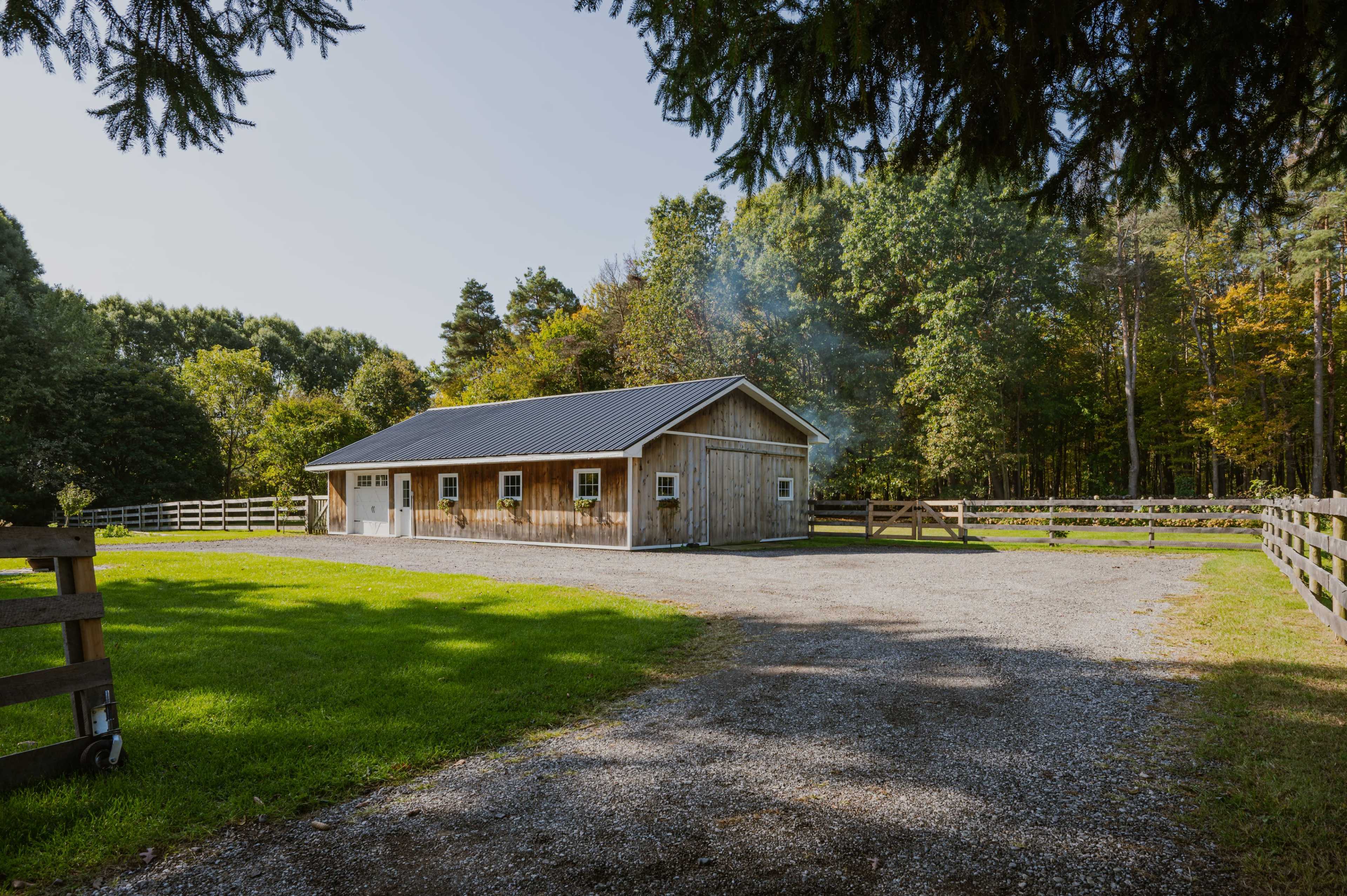 A wooden barn sits beside a gravel path in a green field, surrounded by trees and a wooden fence.