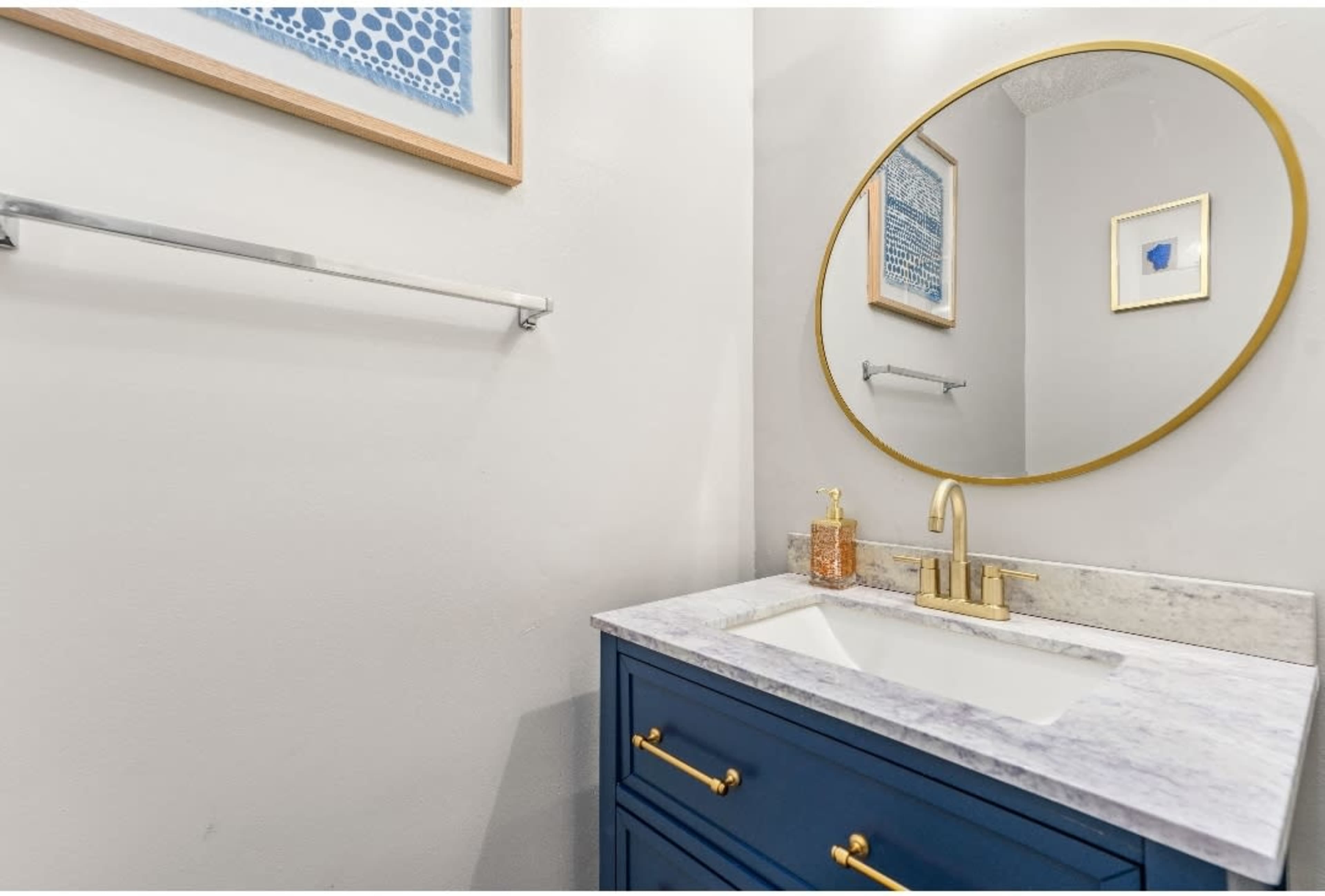 A bathroom vanity featuring a blue cabinet, a marble countertop, a round mirror with a gold frame, and wall art in light blues.