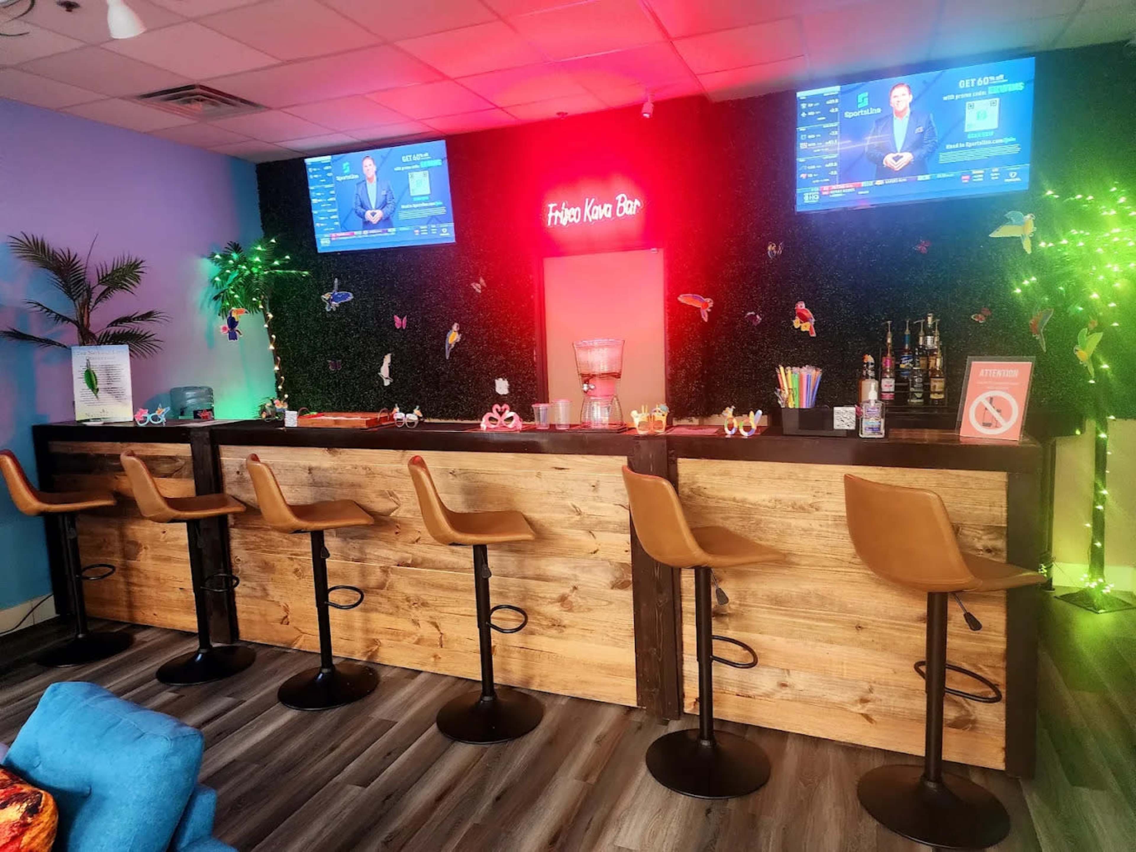 The image shows a bar area with a wooden counter, several high stools, and two televisions displaying news, set against a vibrant green backdrop with decorative plants and lighting.