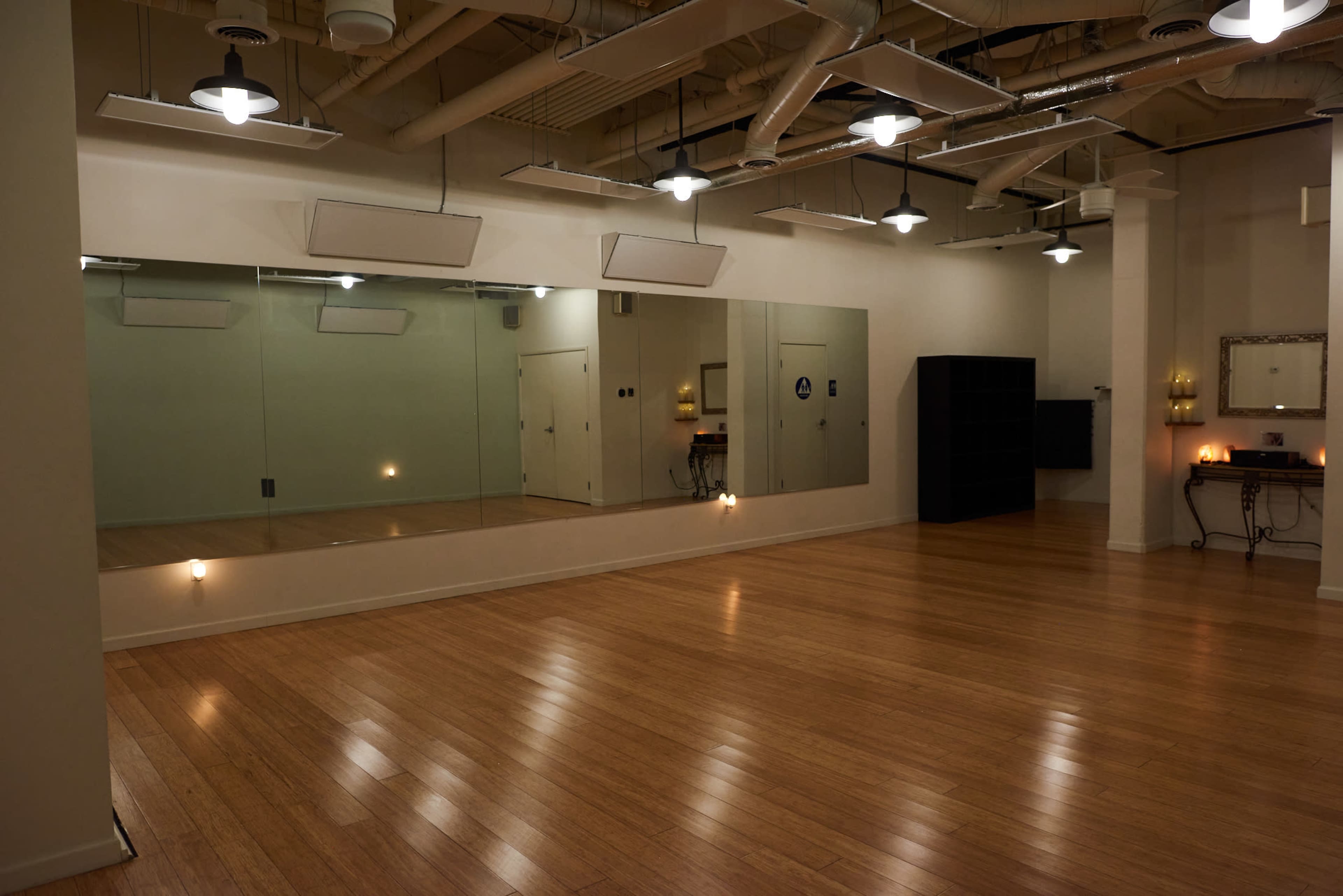 An empty studio space with wooden flooring, large mirrors on one wall, and a few lights hanging from the ceiling.