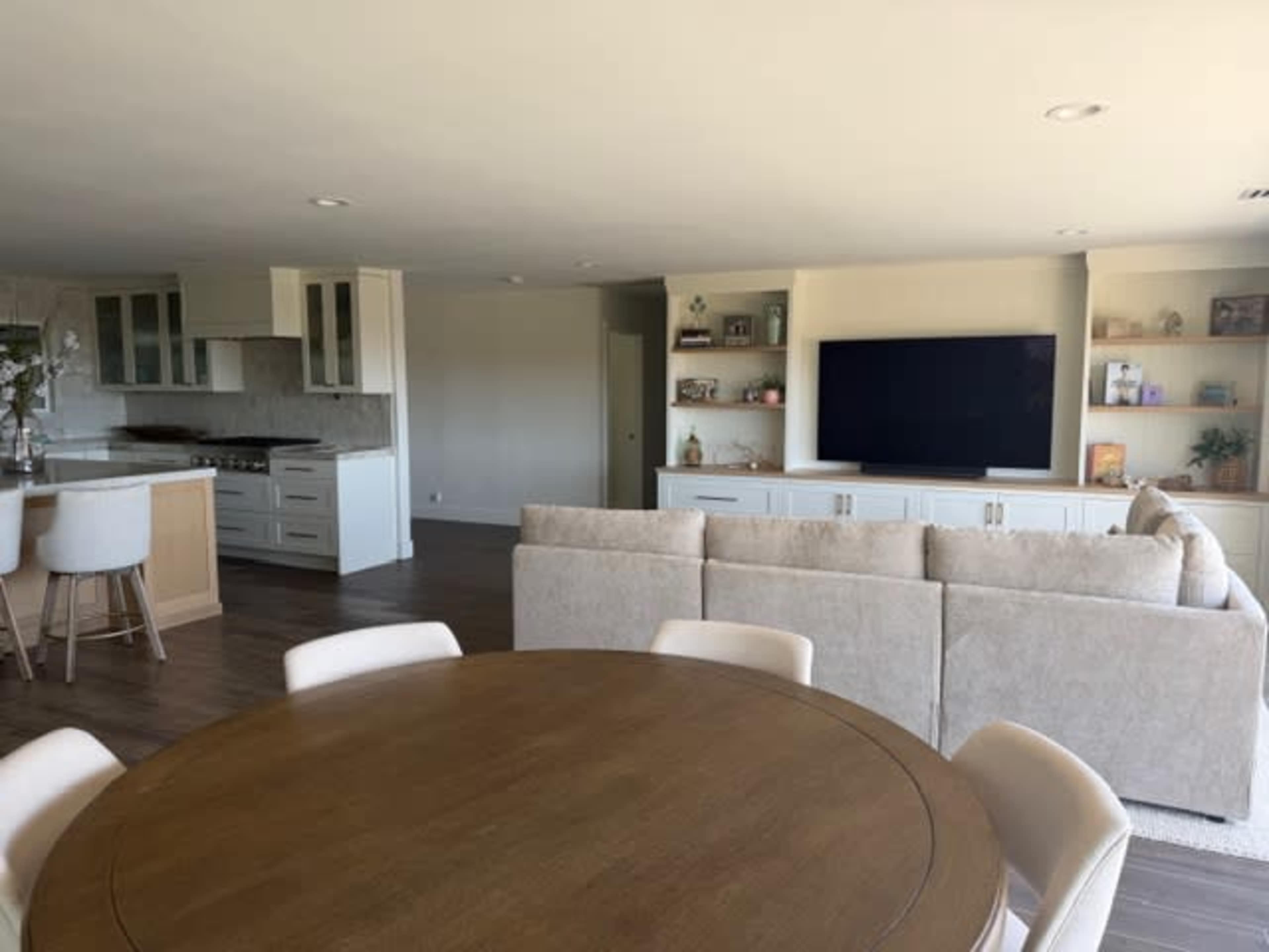 A modern open-concept living space features a circular dining table, a beige sectional sofa, and a wall-mounted television beside a kitchen with light cabinetry.