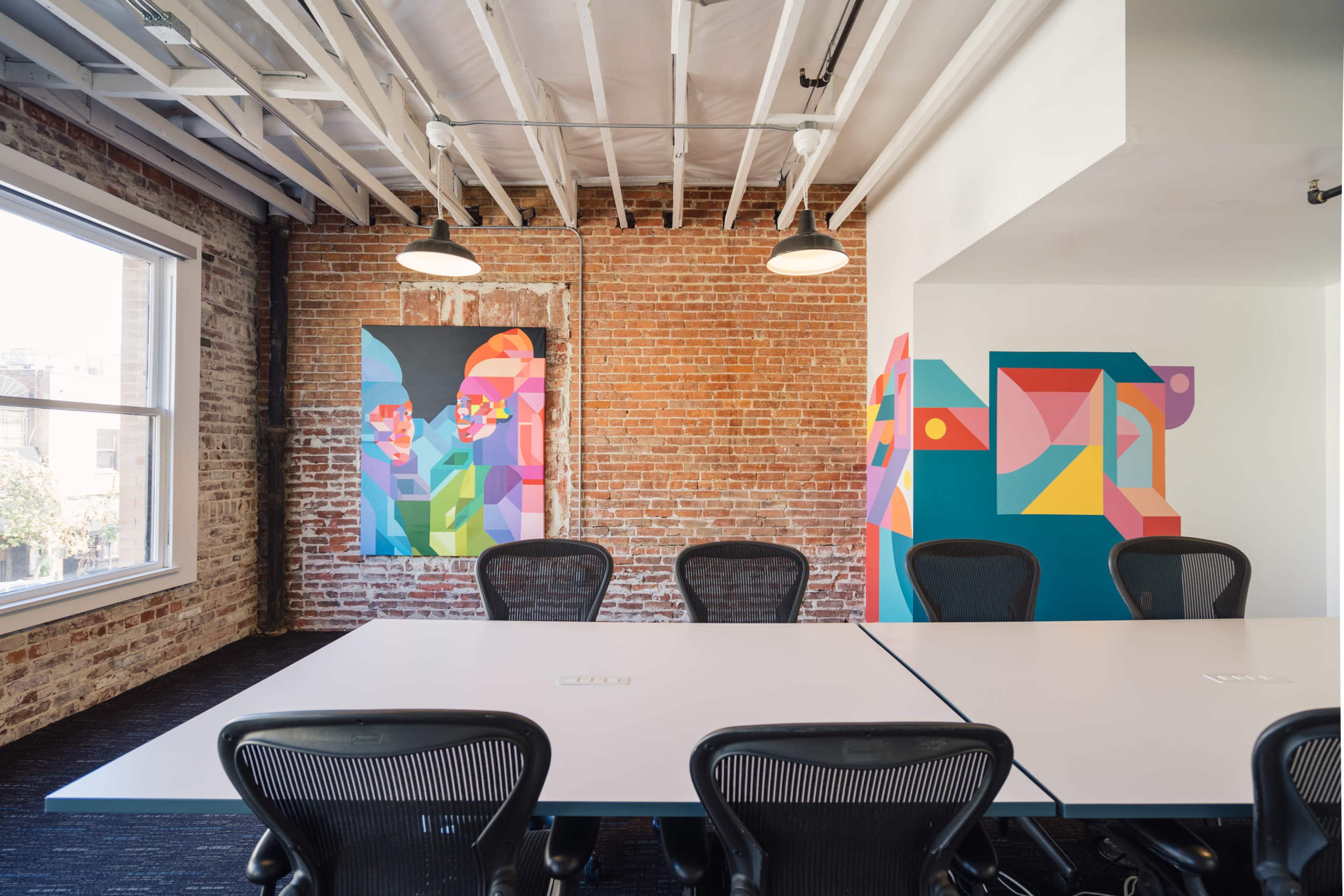 The image shows a modern office space with exposed brick walls, a long white table, and colorful abstract artwork on the walls.