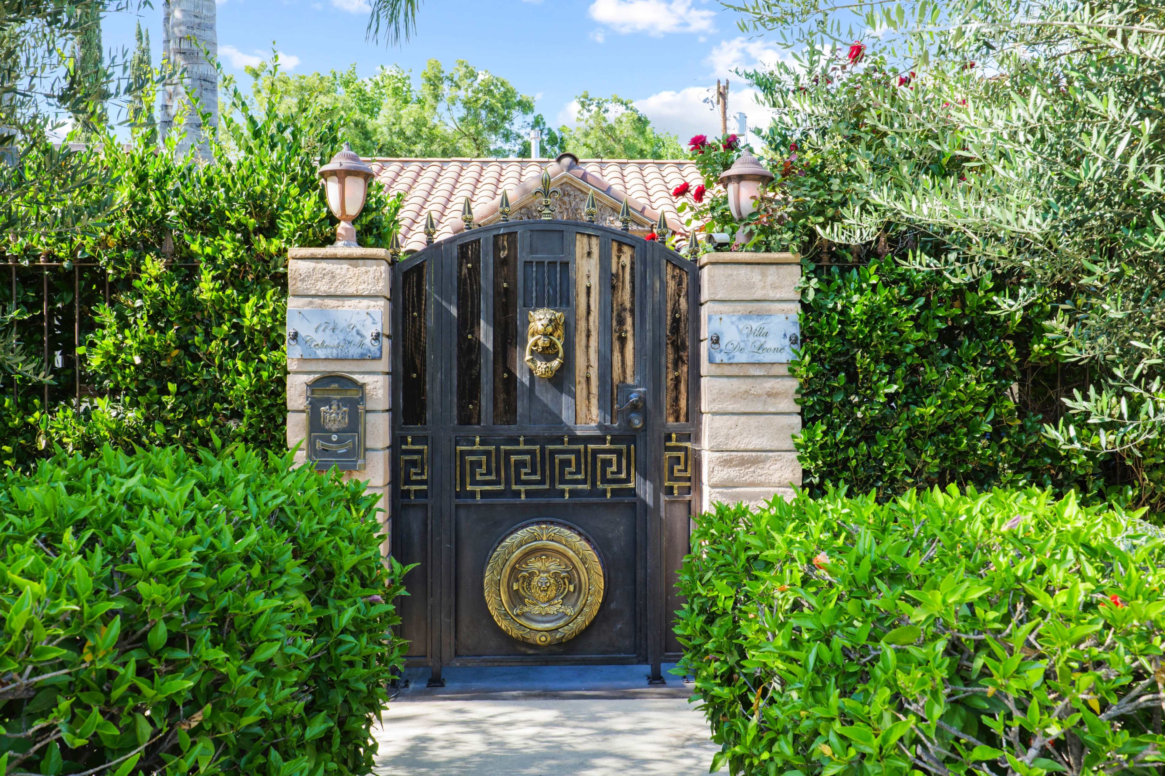 A metal gate with a lion's head knocker and decorative elements is flanked by lush greenery and a cobblestone pathway.