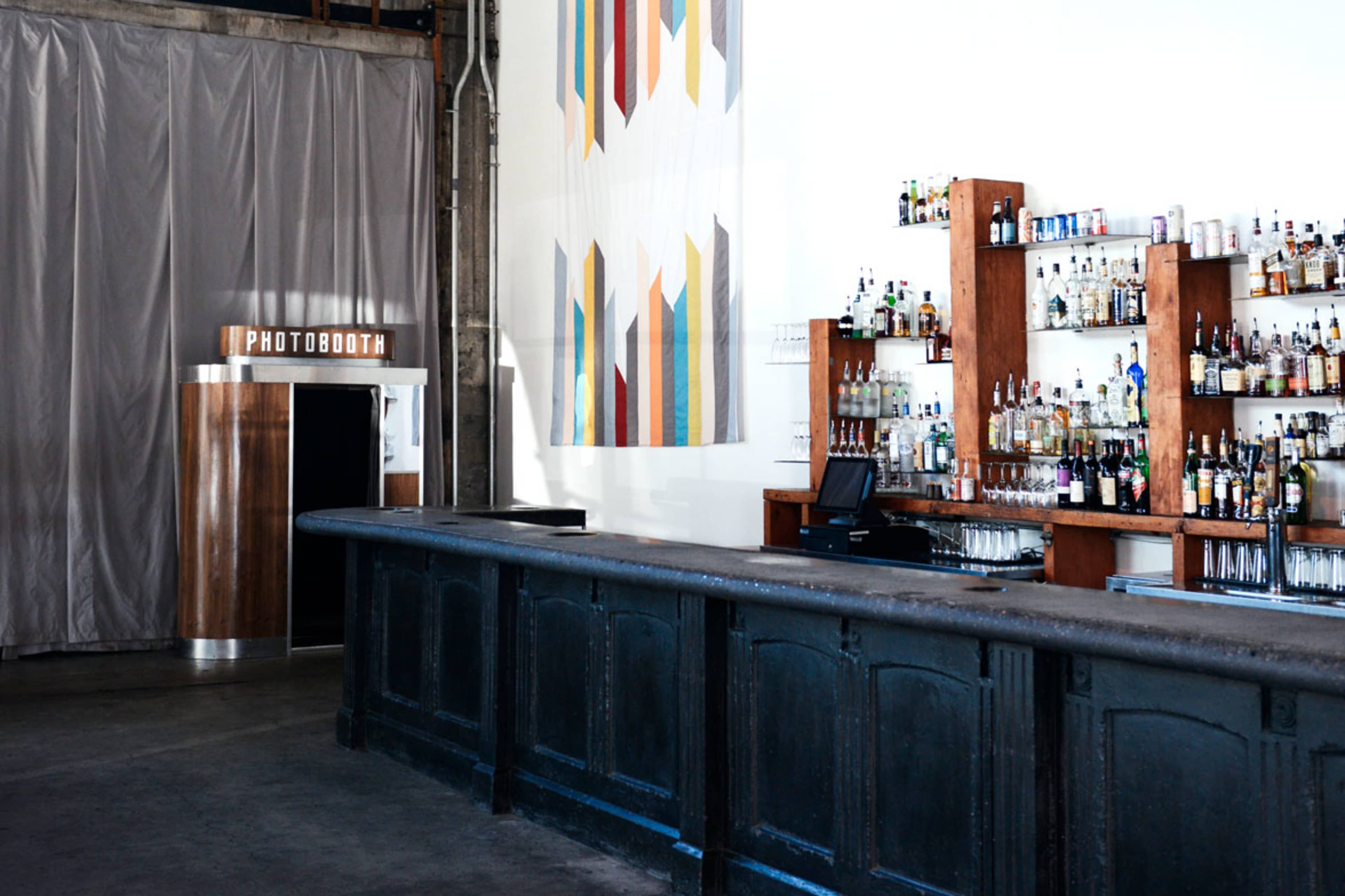 A bar area with a long, dark countertop, a shelf filled with various bottles in the background, and a photo booth to the left.