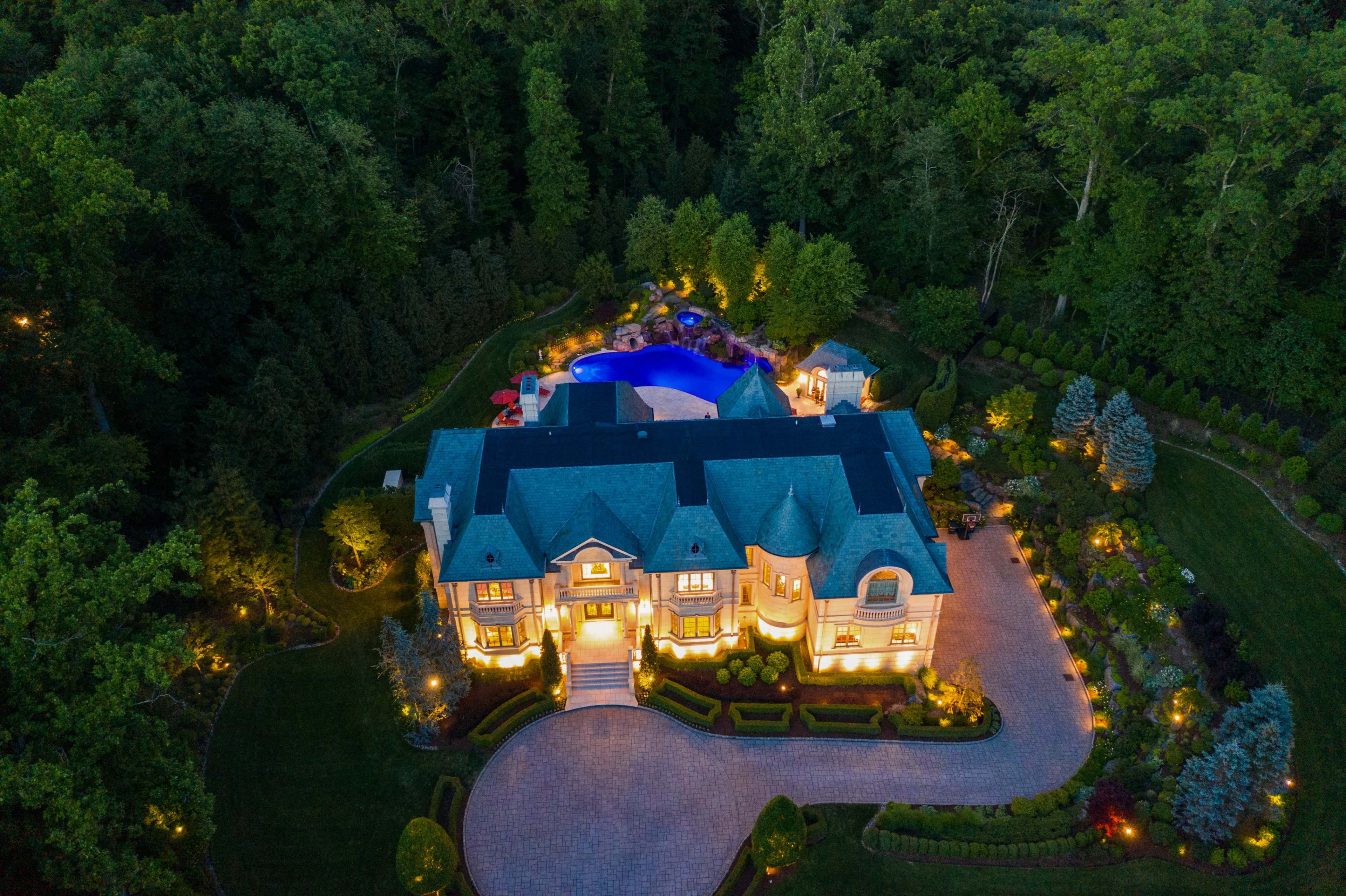 An aerial view of a large, illuminated mansion surrounded by lush greenery and a backyard pool area.