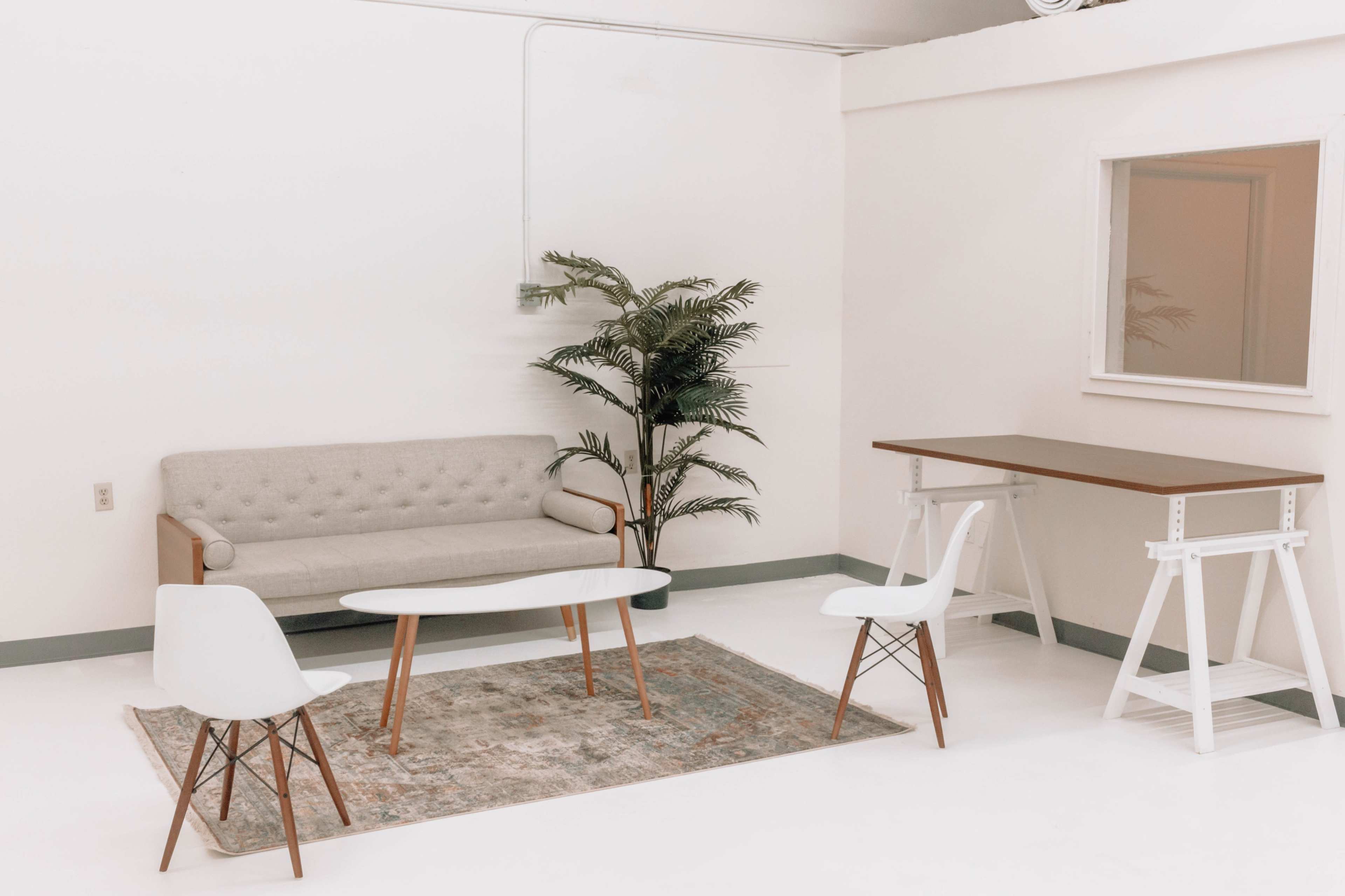 A minimalist interior with a light gray sofa, a small round glass coffee table, two white chairs, a rug, and a potted plant beside a desk against white walls.