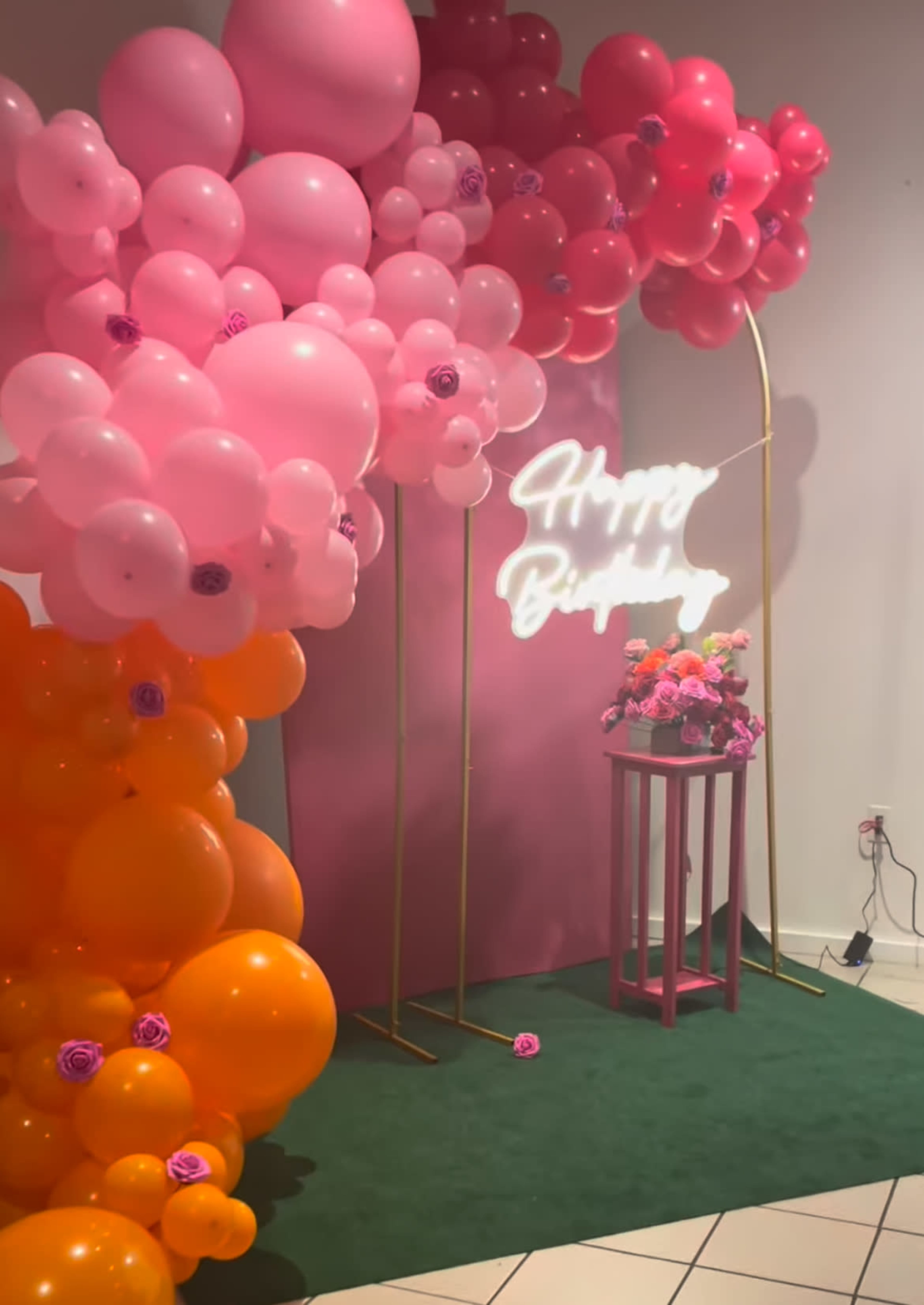 A birthday celebration backdrop featuring clusters of pink, orange, and peach balloons with a neon sign that reads "Happy Birthday" and a small table adorned with flowers.