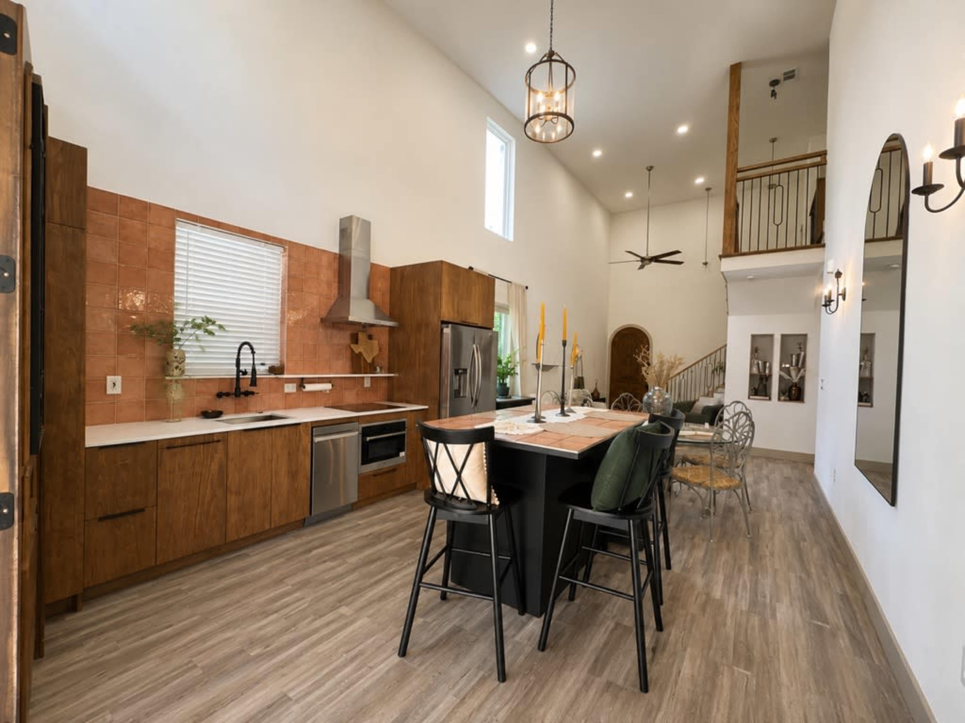 The image shows a modern kitchen with a central island, wooden cabinetry, a stainless steel refrigerator, and large windows allowing natural light.