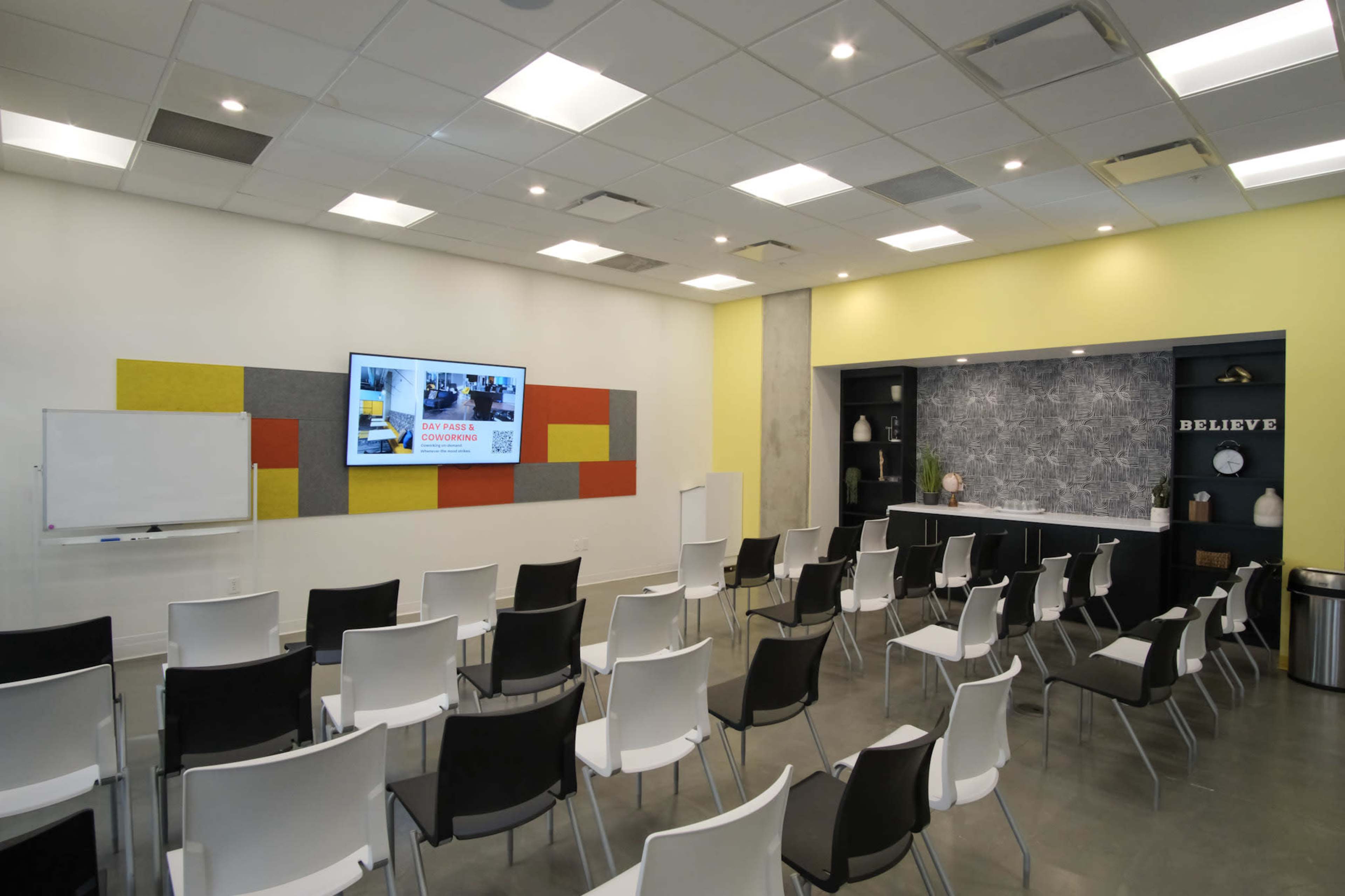 The image shows a modern conference room with rows of white chairs, a large screen displaying information, and a yellow accent wall featuring decorative elements.