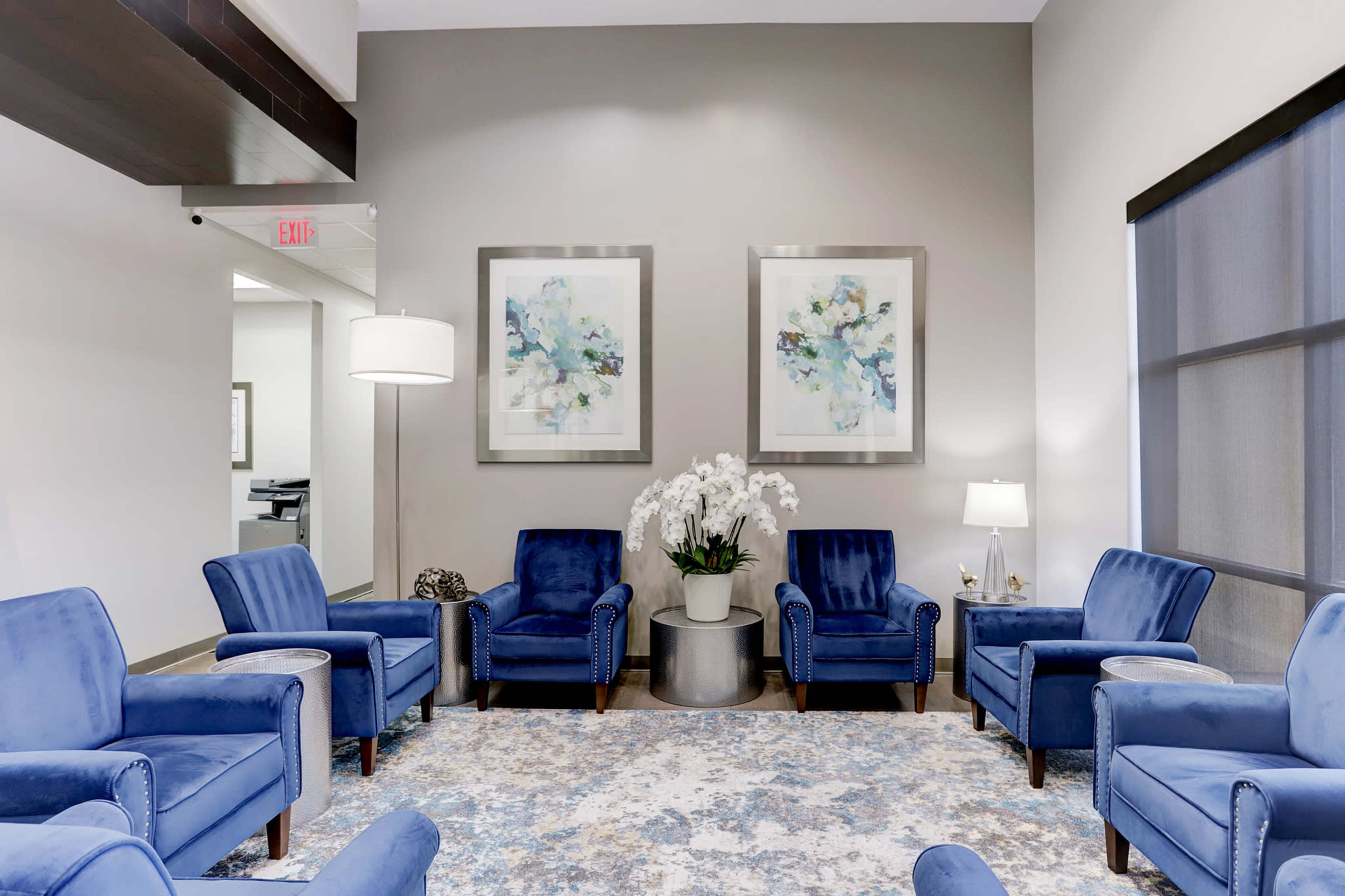 The image shows a waiting area with blue upholstered chairs arranged around a round table, featuring two abstract paintings on the wall and a floor lamp.