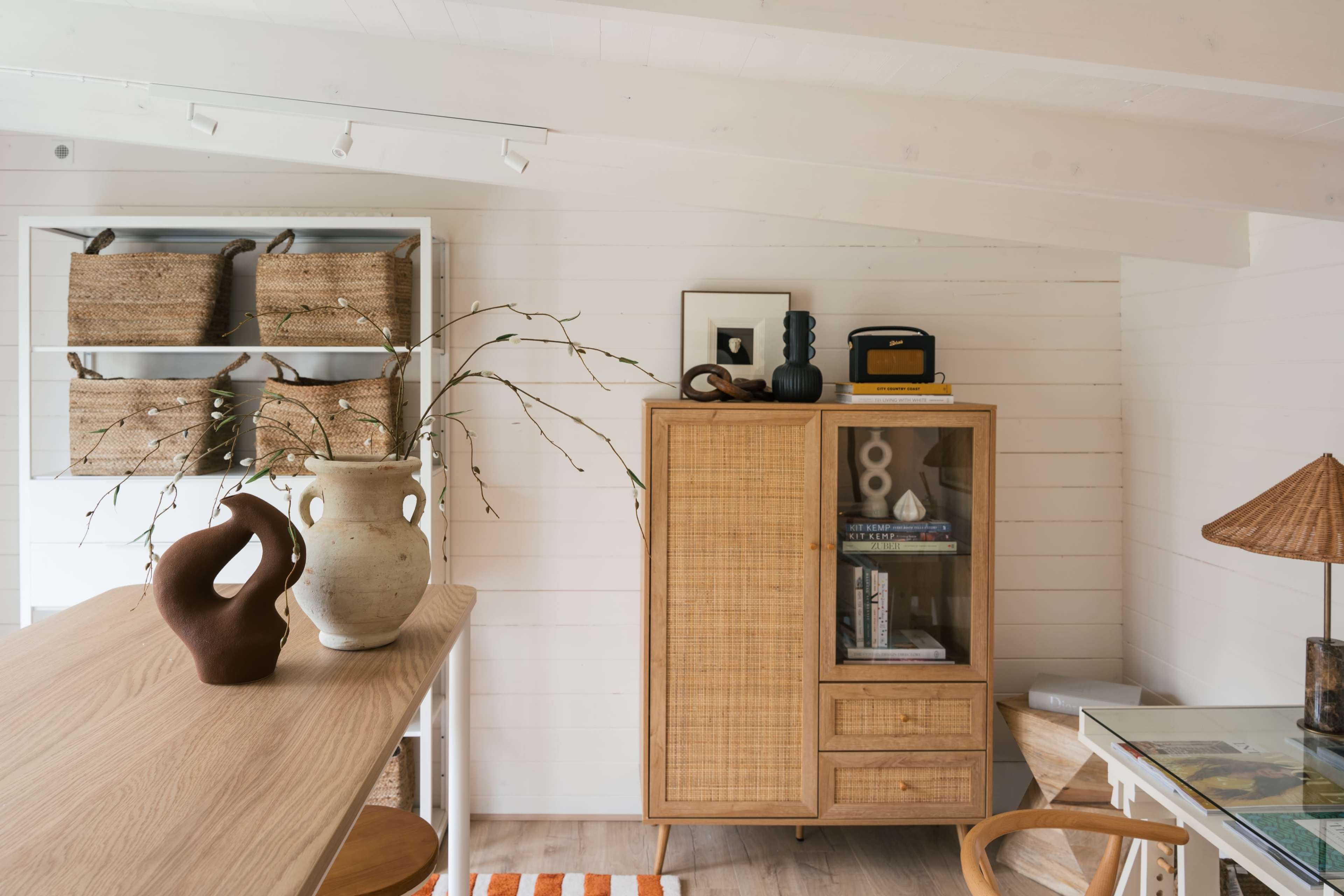 A cozy interior with a wooden table, a rattan cabinet, and decorative elements including a vase and woven wall baskets.