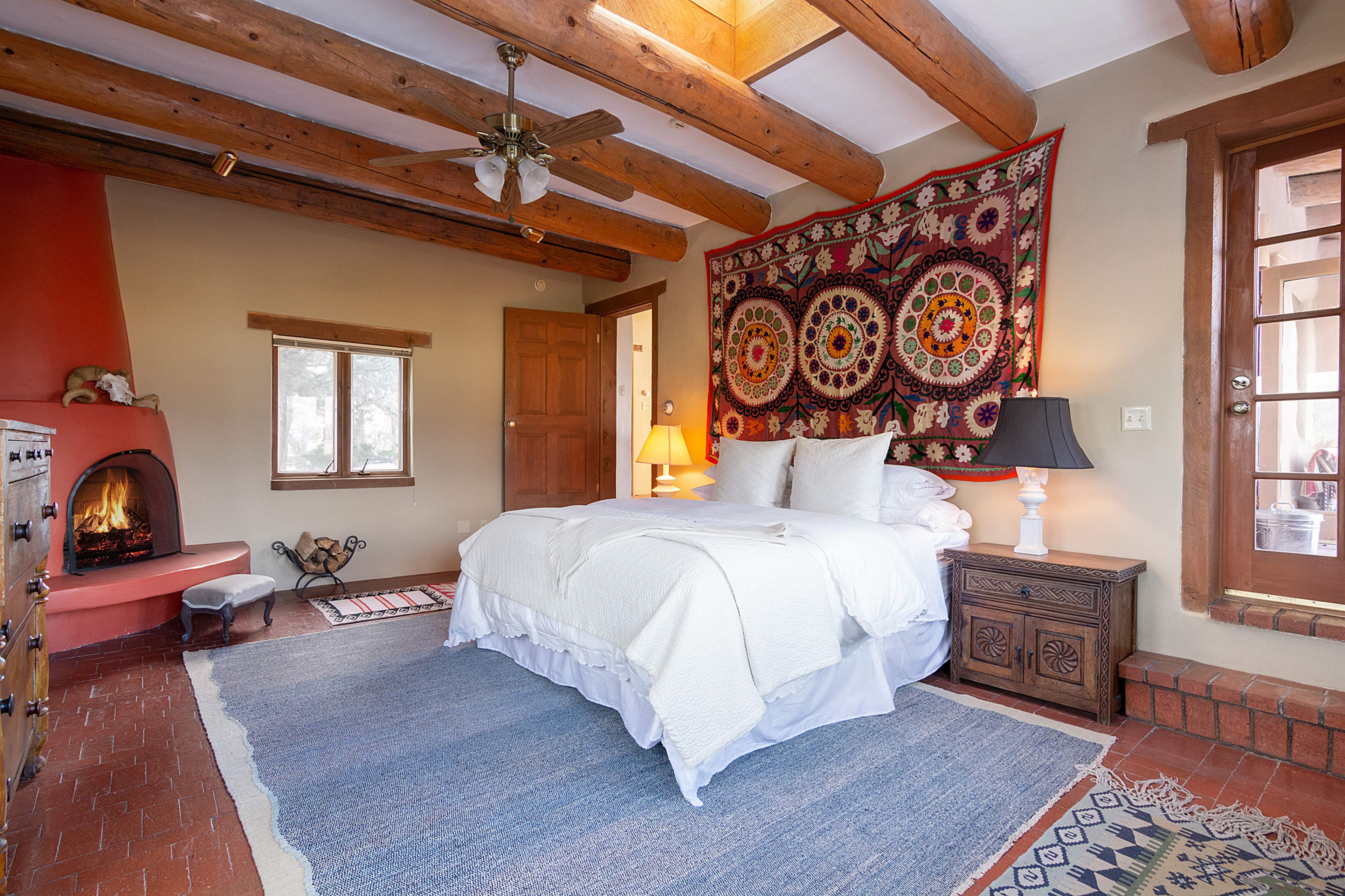 A cozy bedroom features a bed with a white coverlet, a colorful wall tapestry, and a small fireplace, with wooden beams overhead and a door leading outside.