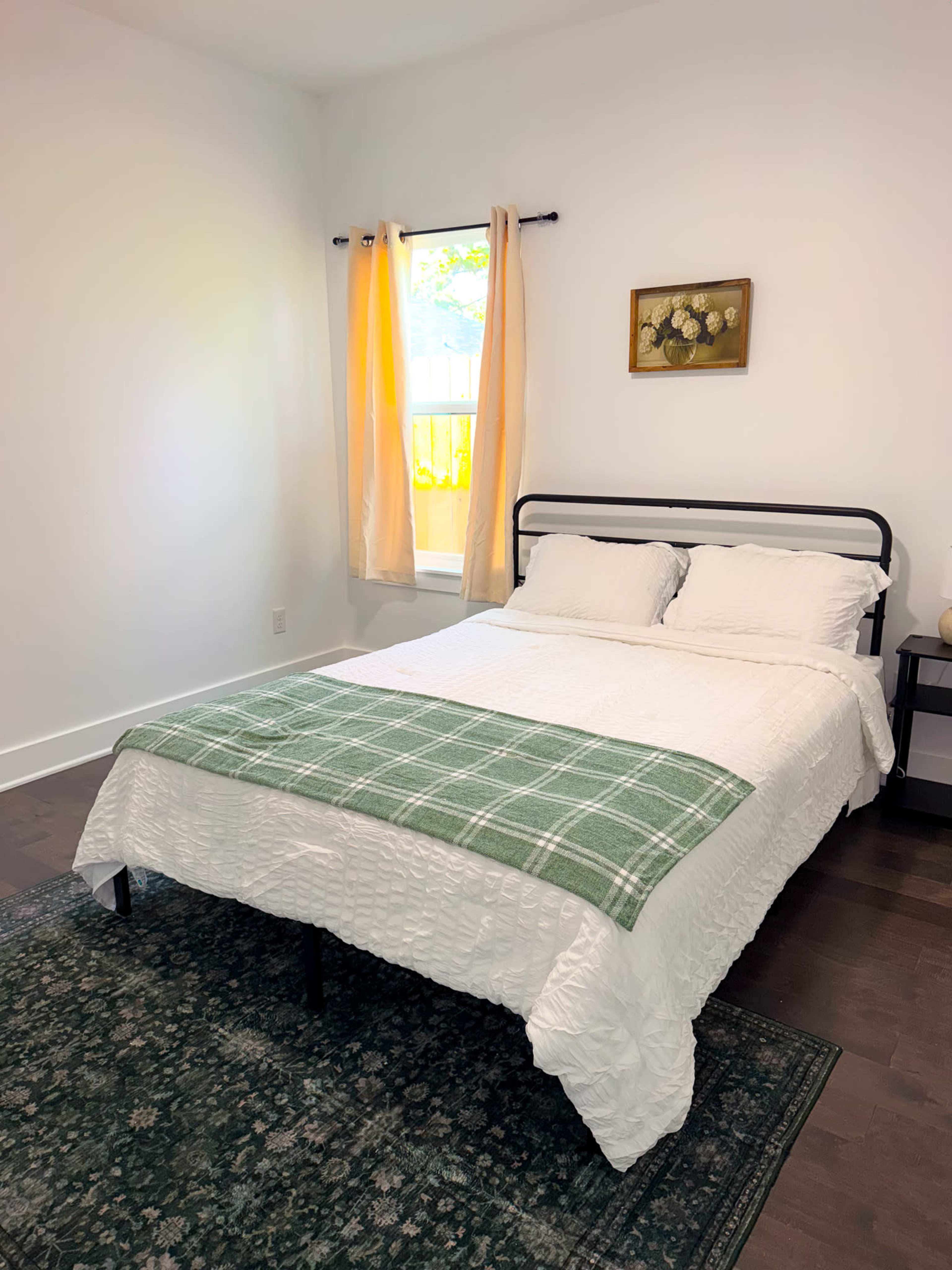 The image shows a neatly made bed with a green plaid throw blanket, situated in a bright, minimalistic bedroom with a window and a small decorative painting on the wall.