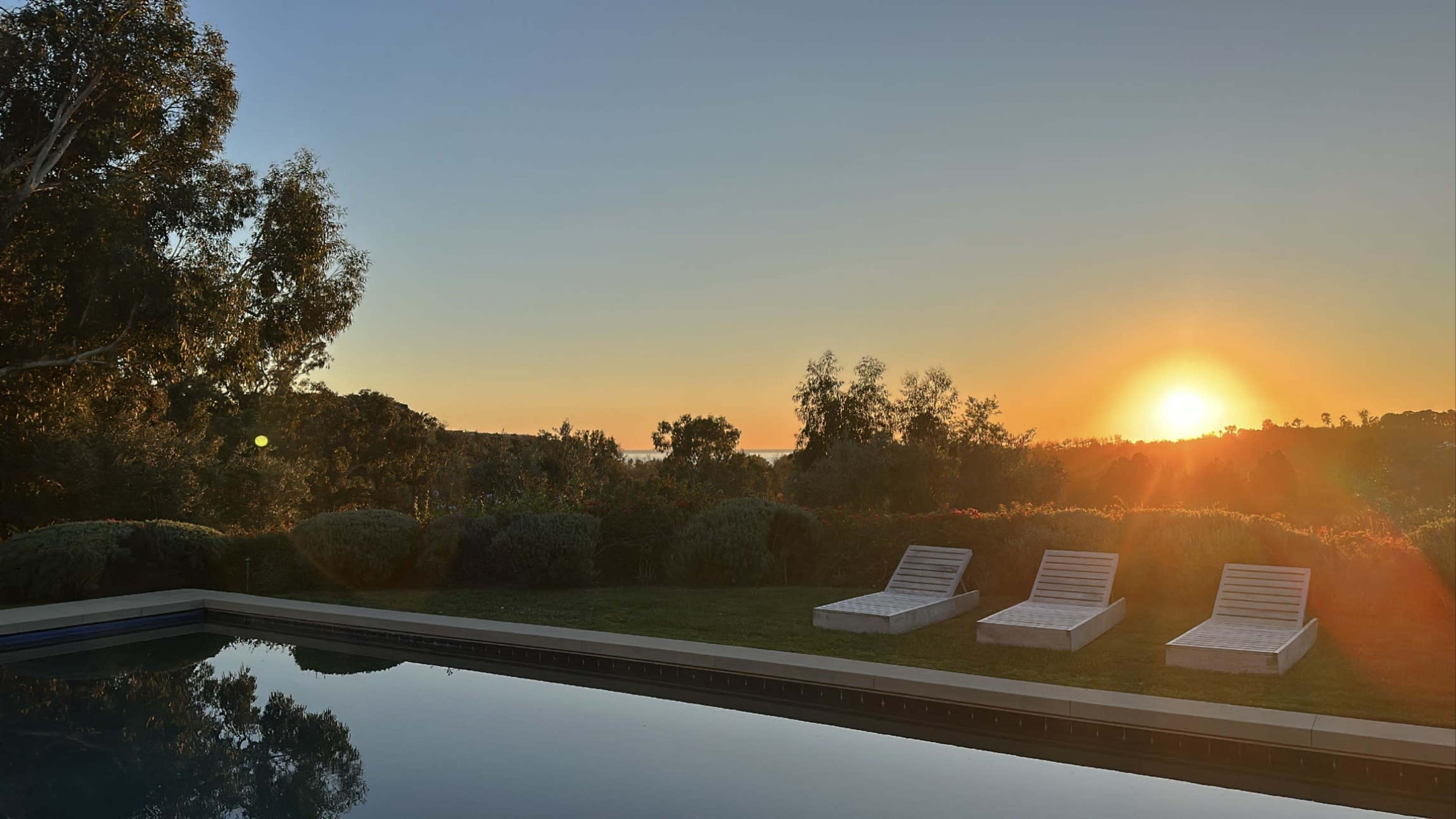 The scene shows a serene outdoor area with three lounge chairs beside a calm swimming pool, framed by lush greenery and a vibrant sunset in the background.