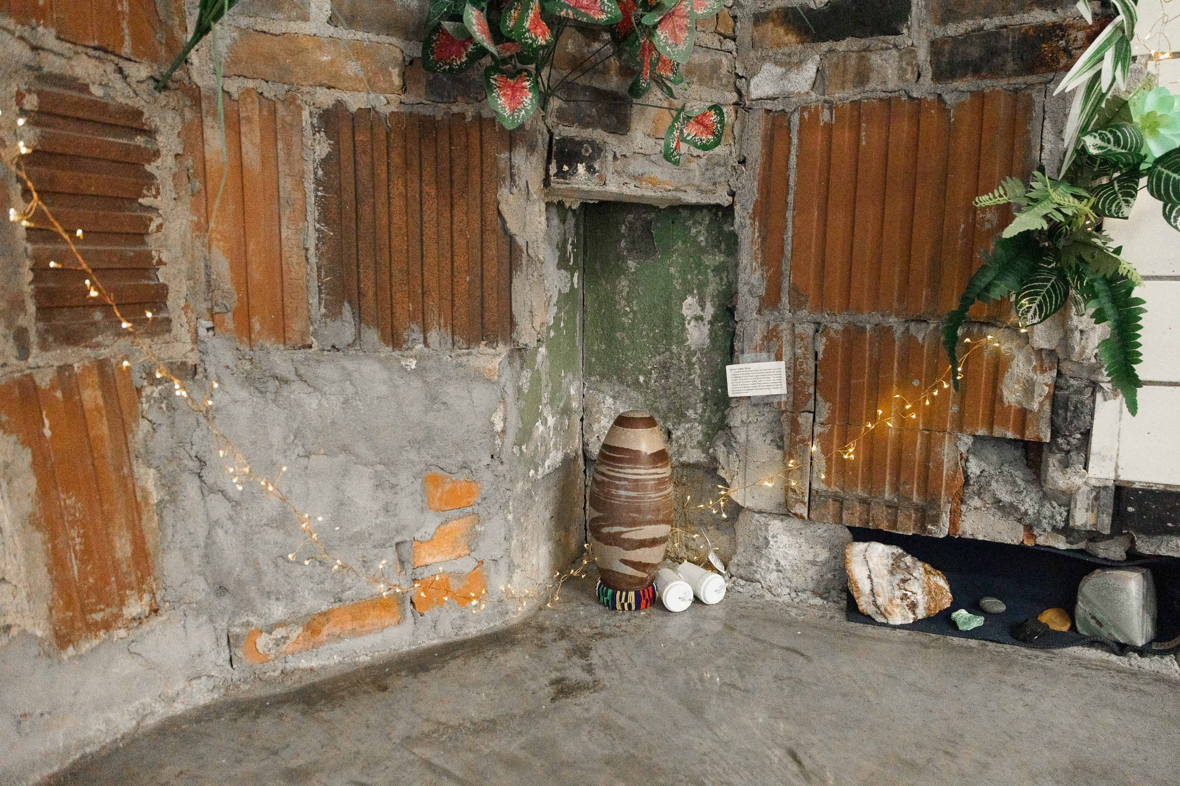 A corner of a room with a textured wall made of bricks and plaster, featuring decorative plants, twinkling lights, and pottery.