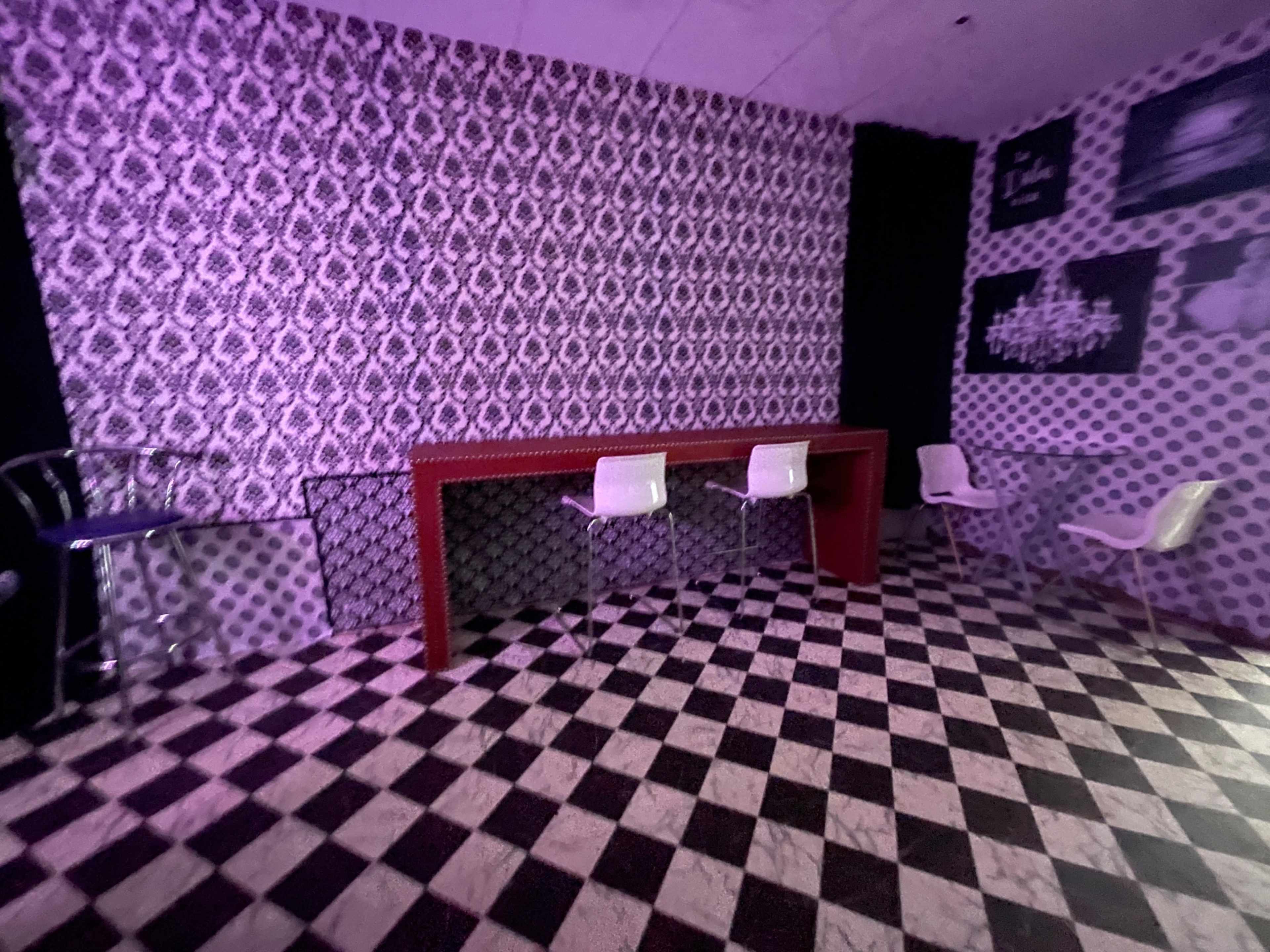 The room features a patterned wallpaper, a red bar table, and white chairs arranged on a black and white checkered floor.