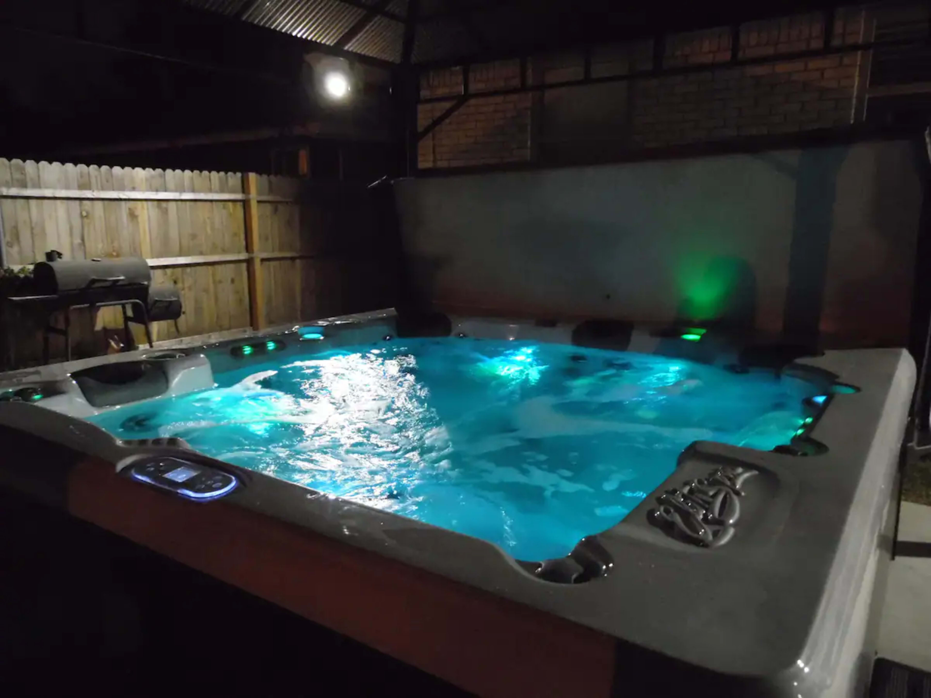 A hot tub with illuminated water is situated under a covered outdoor area at night.