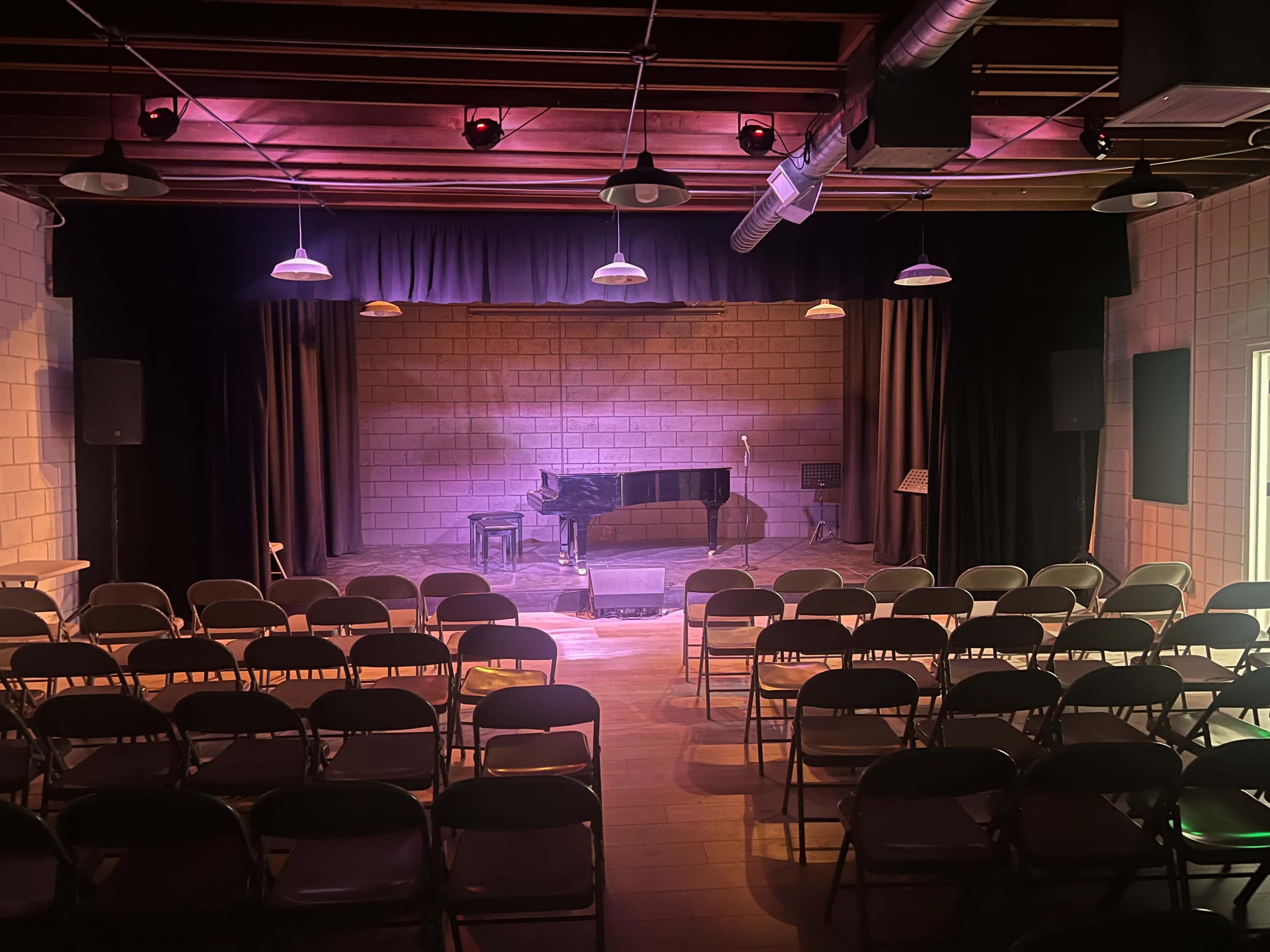 A small theater stage with a grand piano and a row of empty folding chairs facing the stage.
