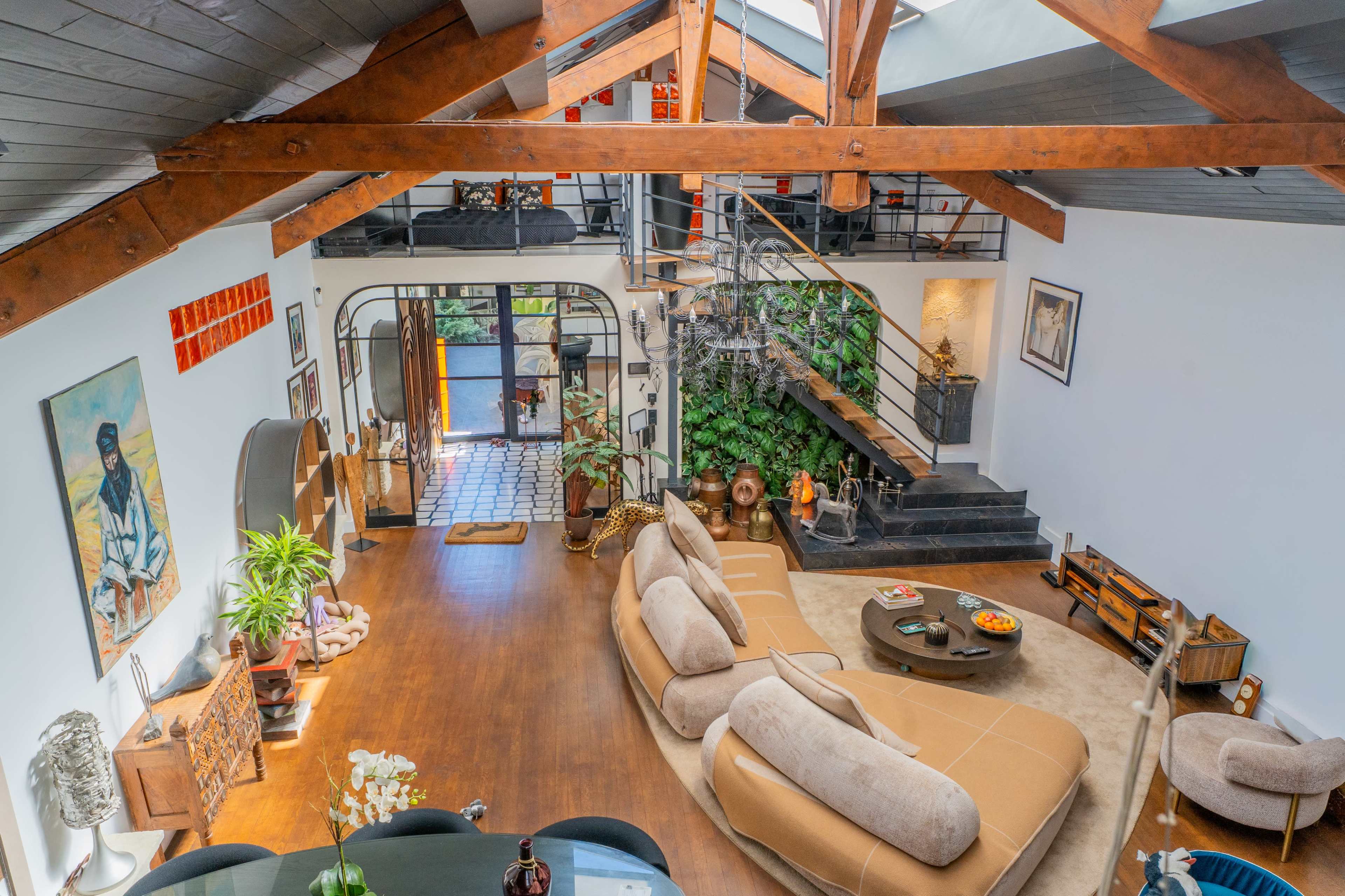 The image shows a spacious, modern living area with high wooden ceilings, large windows, and a staircase leading to an upper level, surrounded by decorative plants and various furniture pieces.