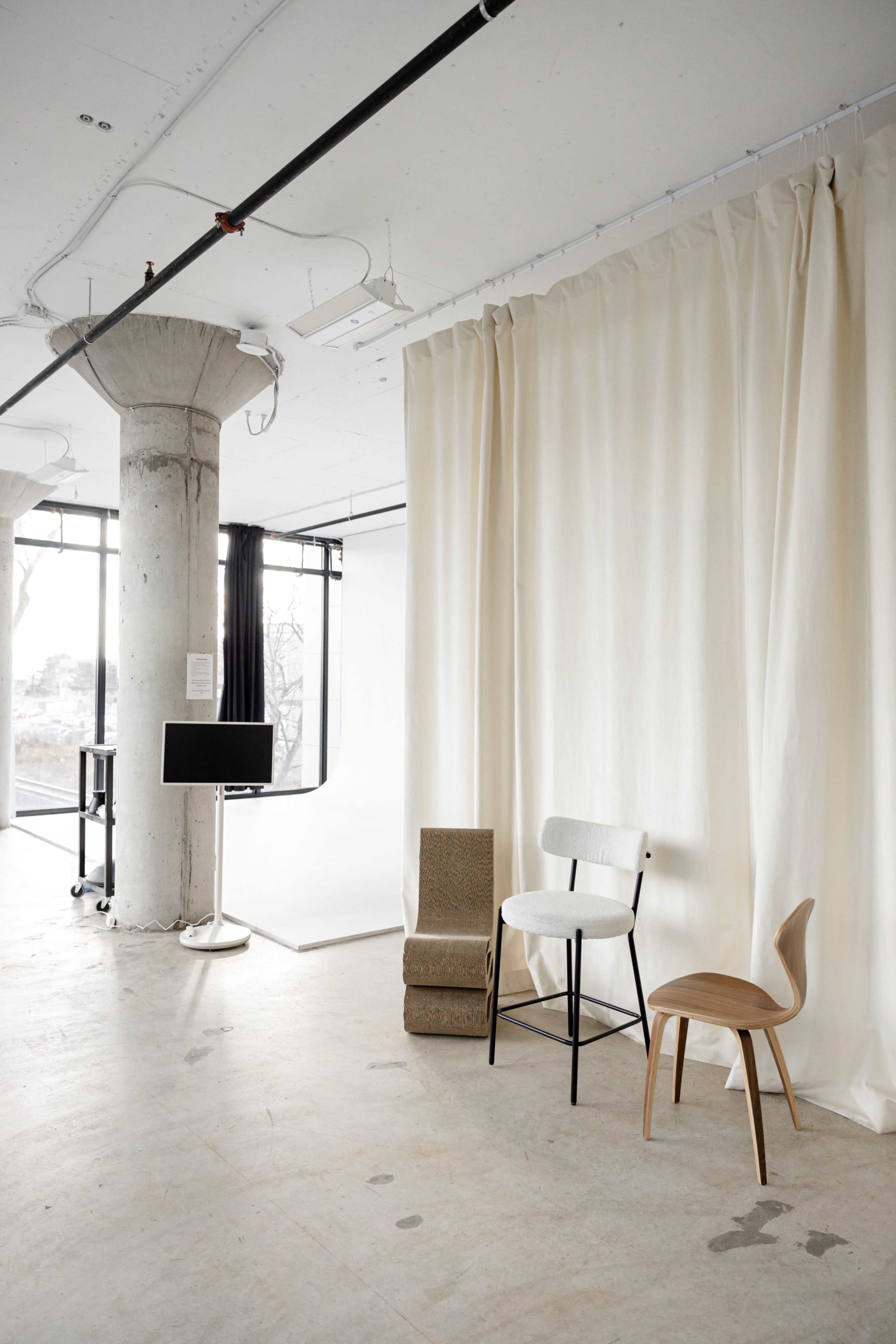 The image shows a minimalist studio space featuring three different chairs, a large curtain, and a concrete pillar, with large windows letting in natural light.