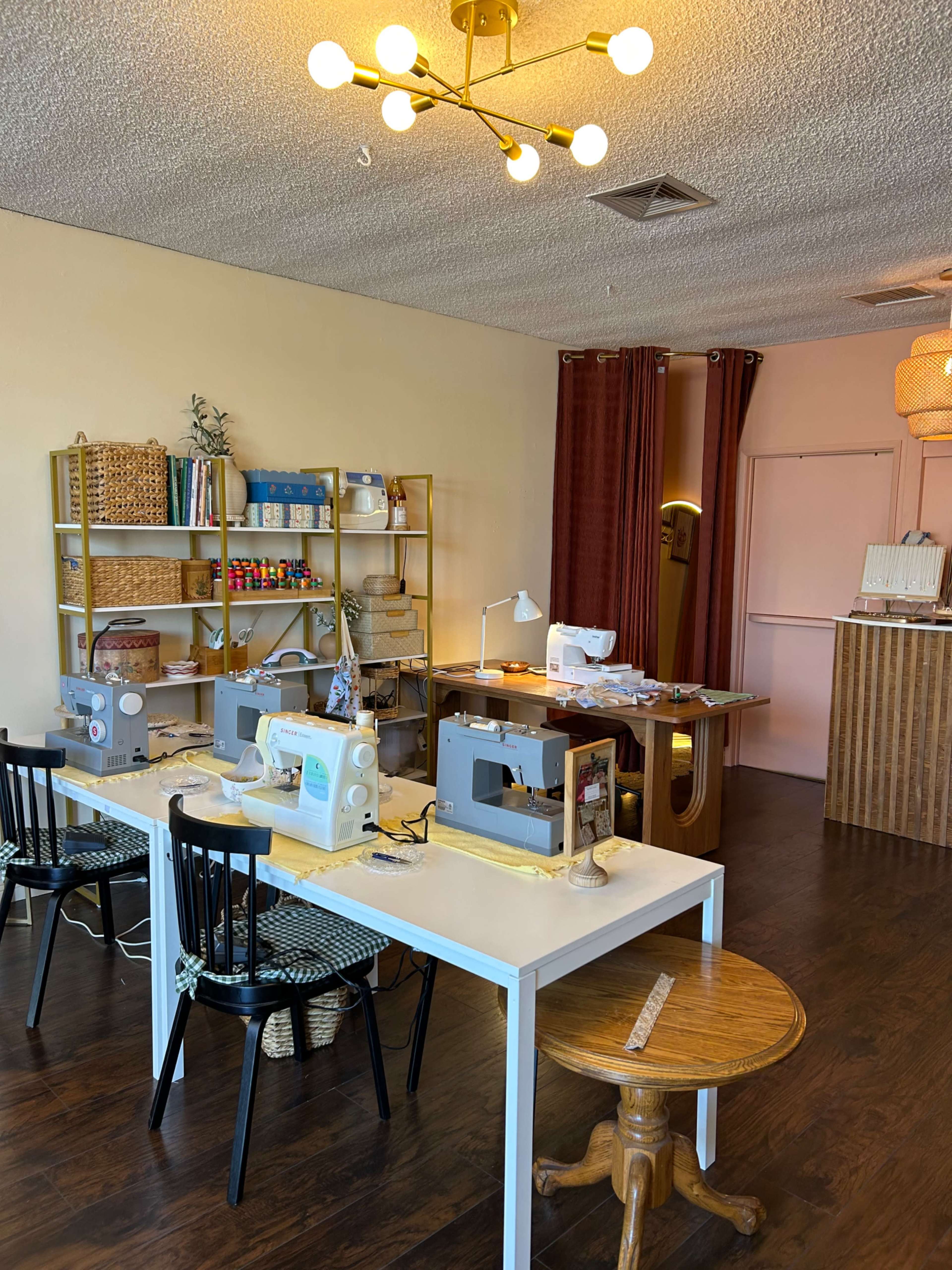 The image shows a crafting room with multiple sewing machines set up on a white table, shelves filled with supplies, and a warm light fixture above.