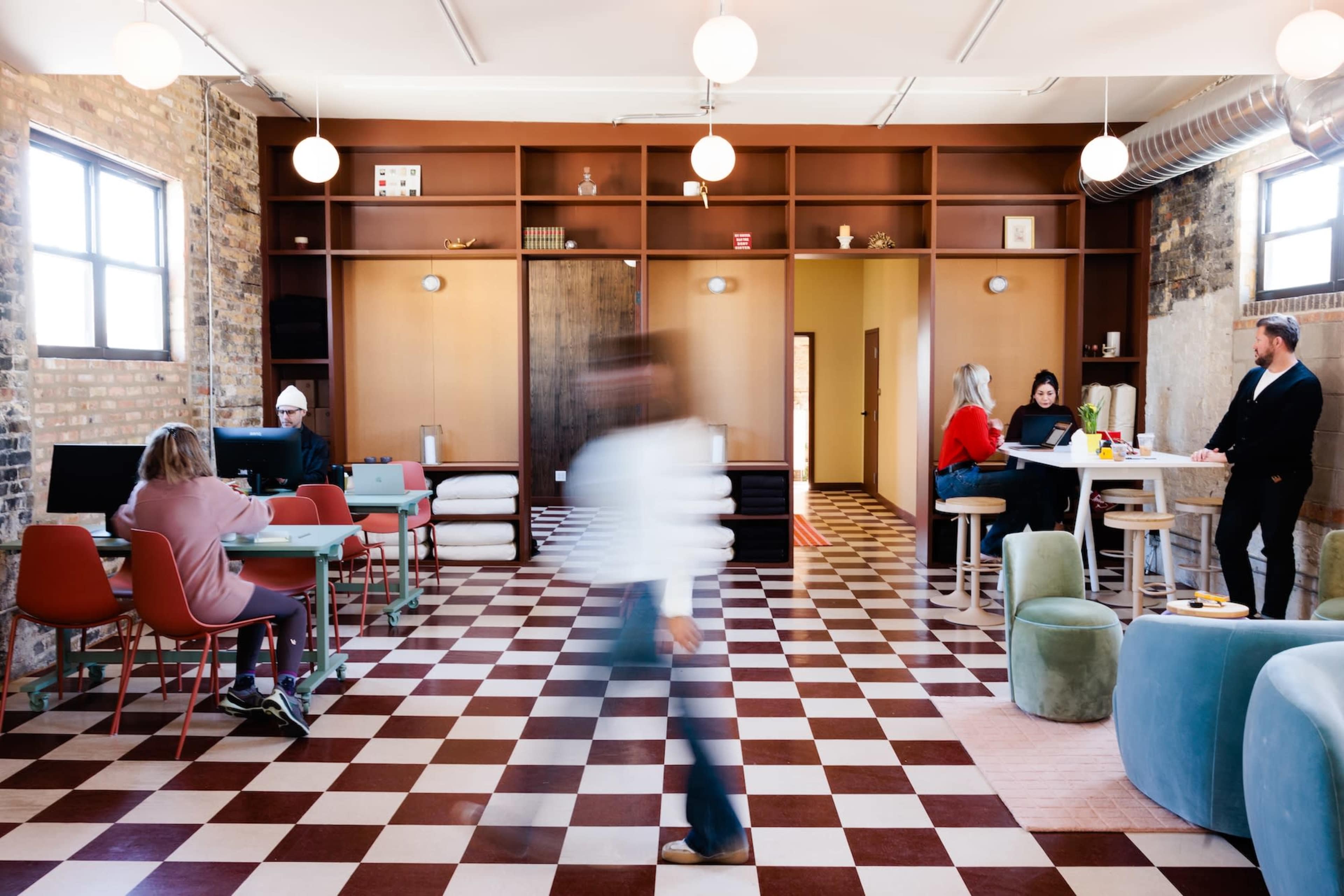 A modern coworking space features a checkered floor, various seating areas, and individuals engaged in work and conversation.