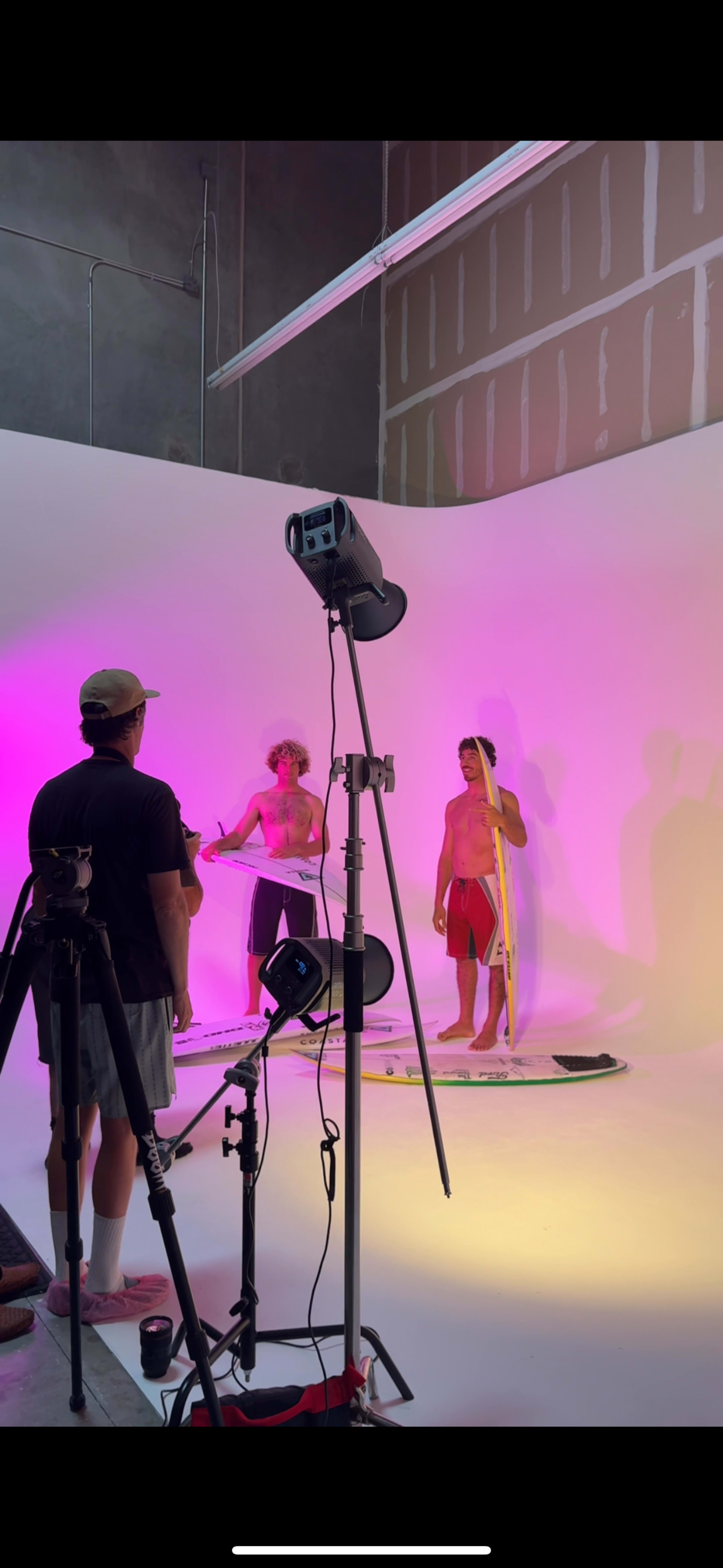 Two shirtless individuals stand on surfboards in a studio with a gradient backdrop illuminated in pink and yellow, while a person in a hat adjusts equipment nearby.