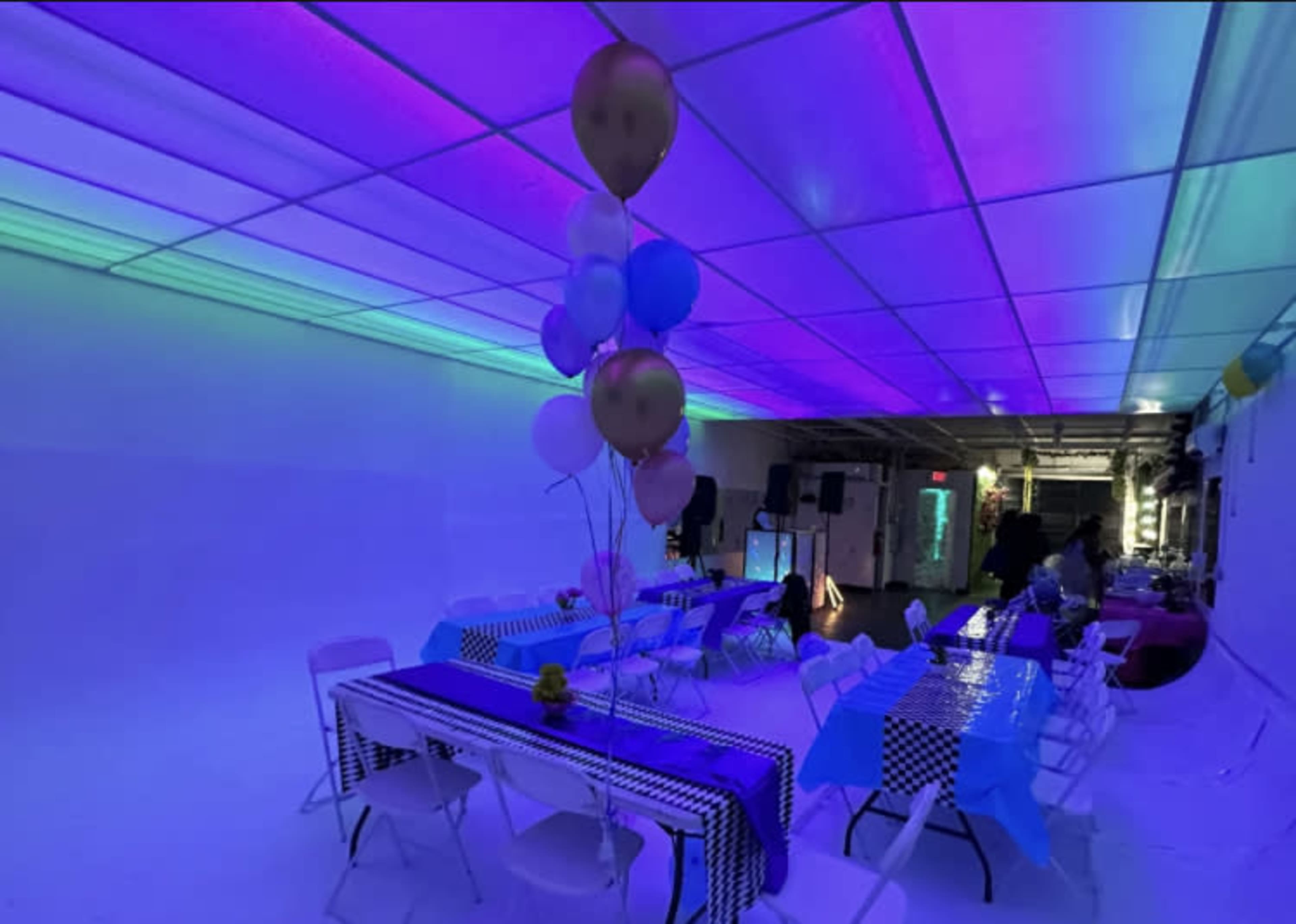 The interior of a brightly lit event space features tables decorated in blue and black, with colorful balloons clustered in the center.