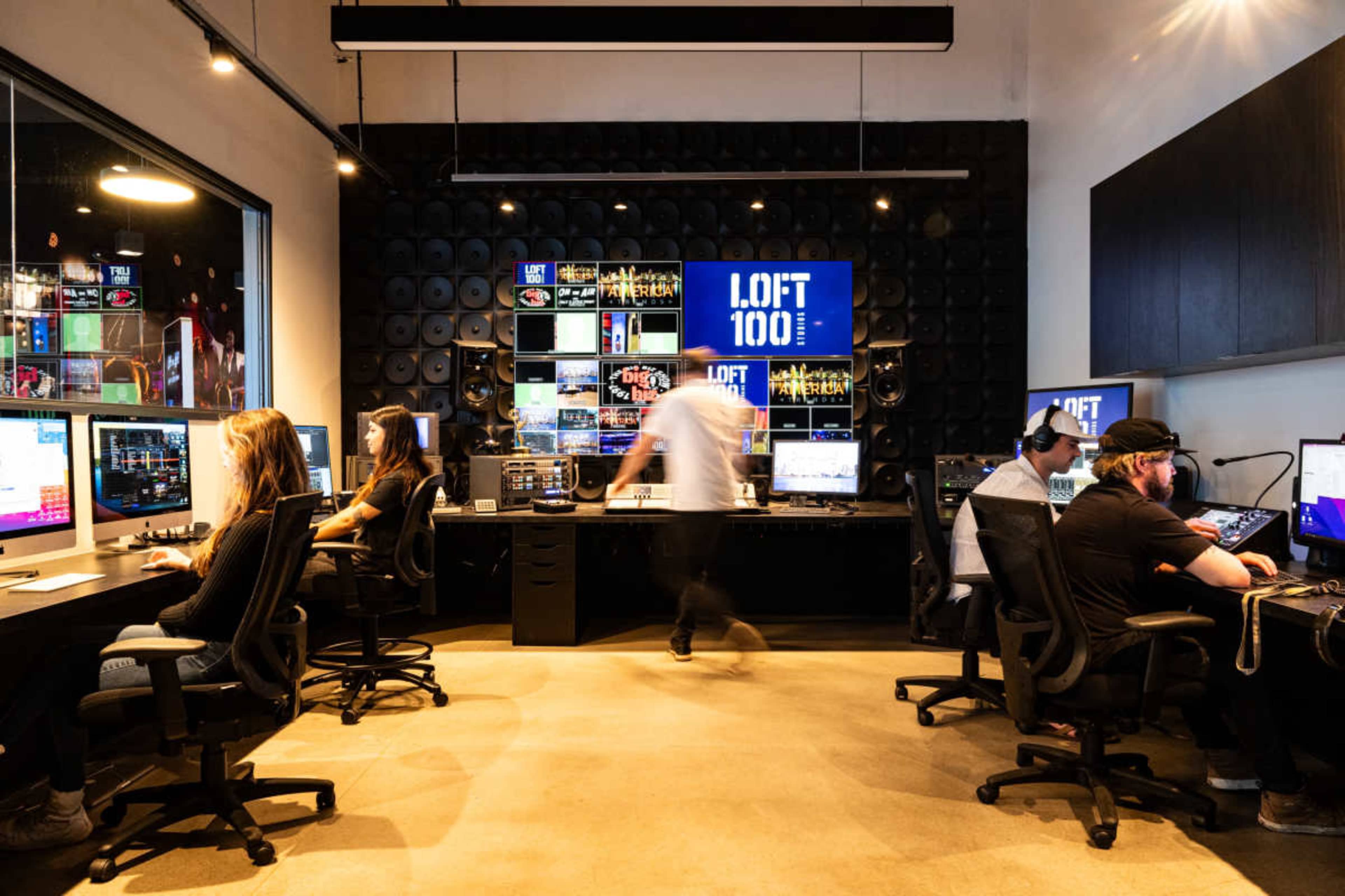 A group of people work at computers in a modern office setup with large screens displaying graphics and sound equipment on the walls.