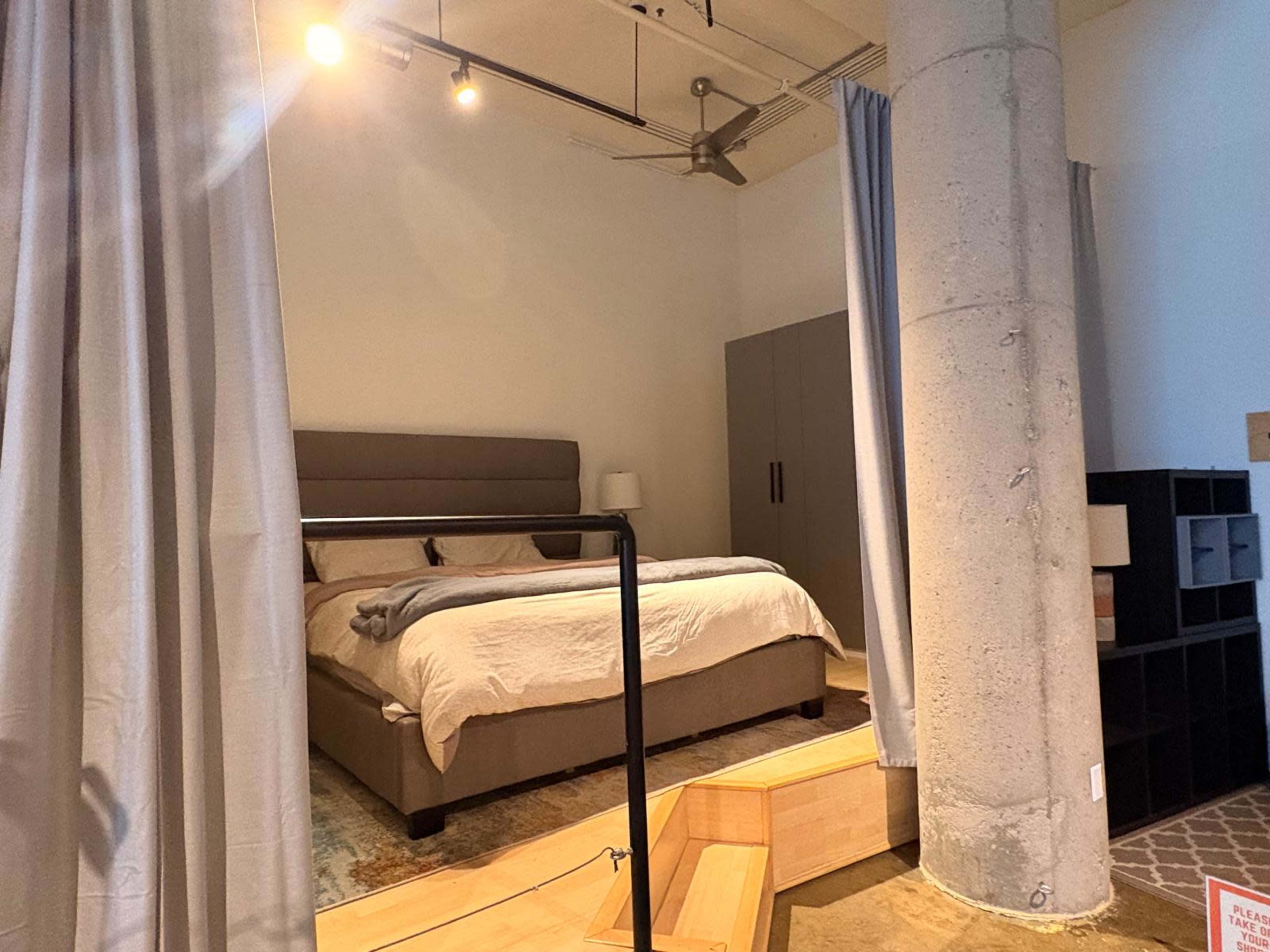 A modern bedroom features a bed with a gray headboard, a nearby wardrobe, and a decorative area rug, divided by curtains for privacy.