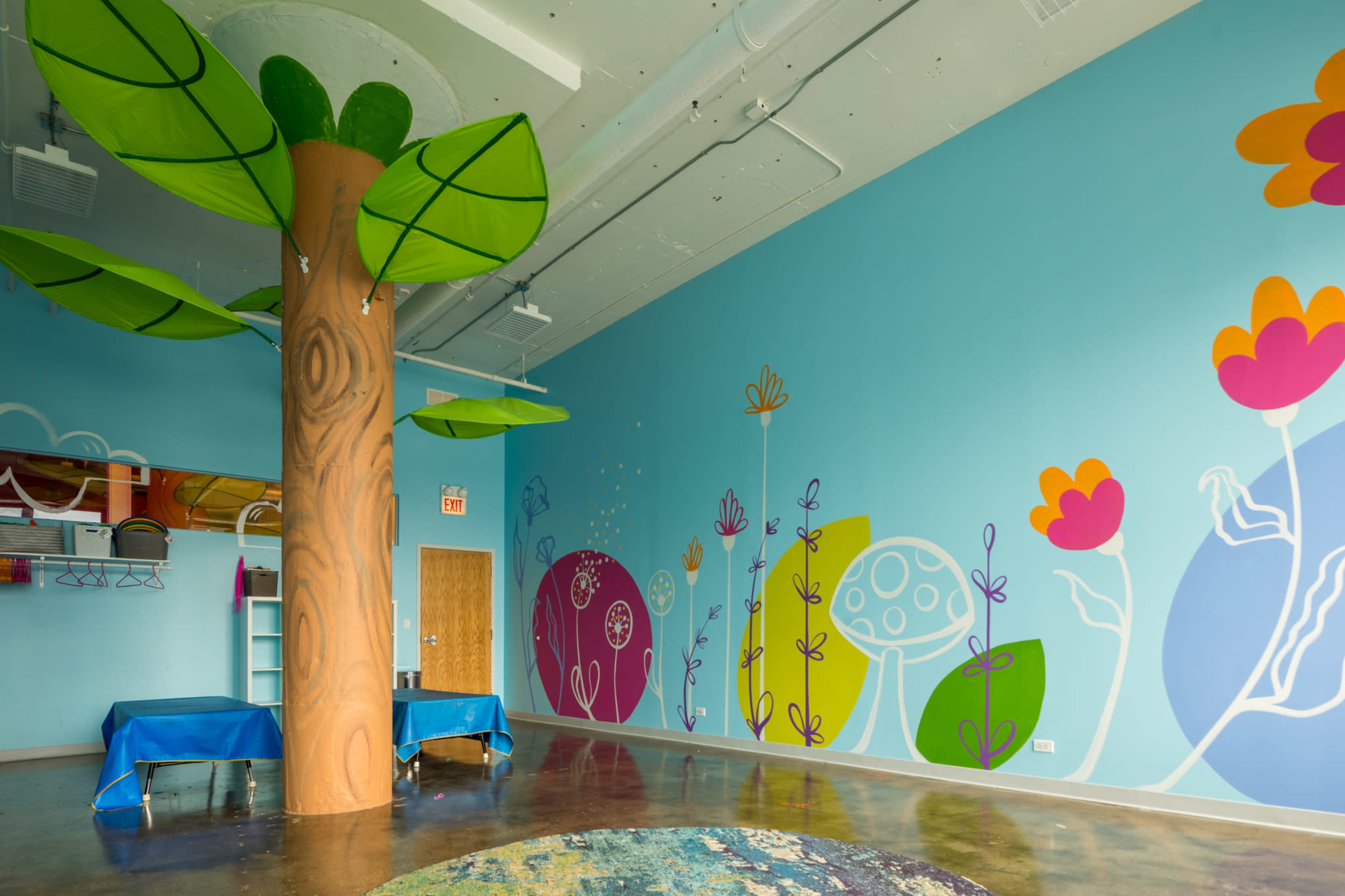 The room features a large tree trunk column with green leaf canopies and colorful, whimsical murals of flowers and shapes on the blue walls.