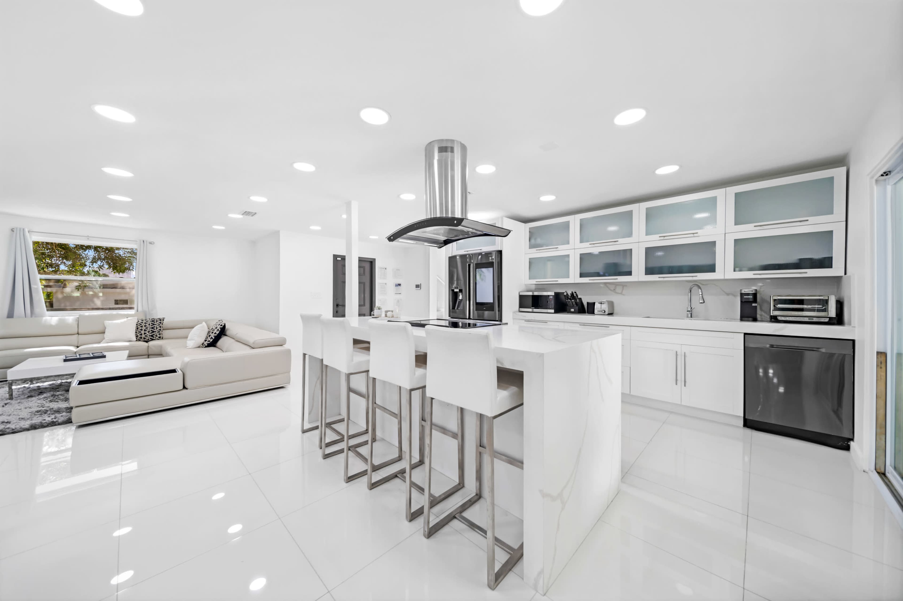The image shows a modern kitchen with white cabinetry, a marble island, and bar stools, adjacent to a living area with a sectional sofa.