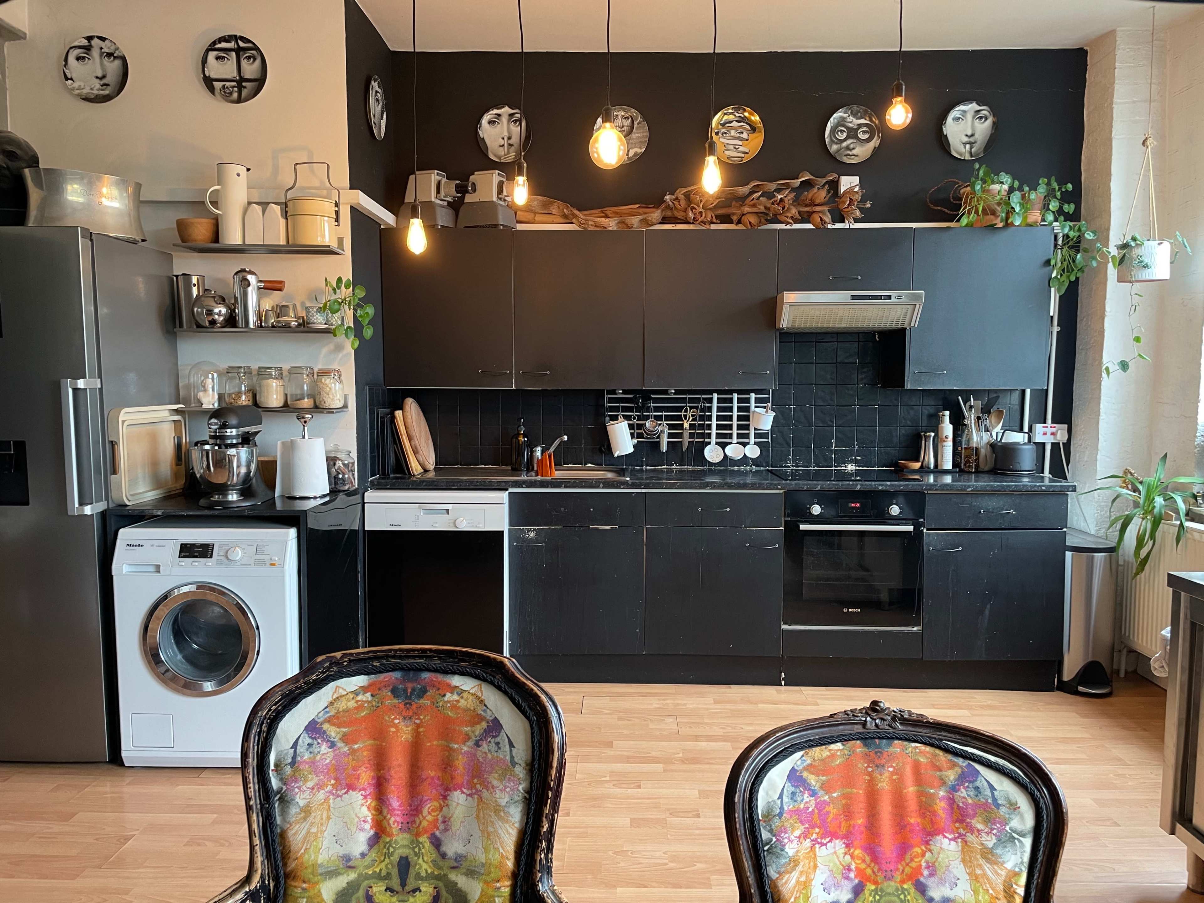 A modern kitchen with black cabinets, a stainless steel refrigerator, and several decorative plates on the wall, alongside a washing machine and two embellished chairs in the foreground.