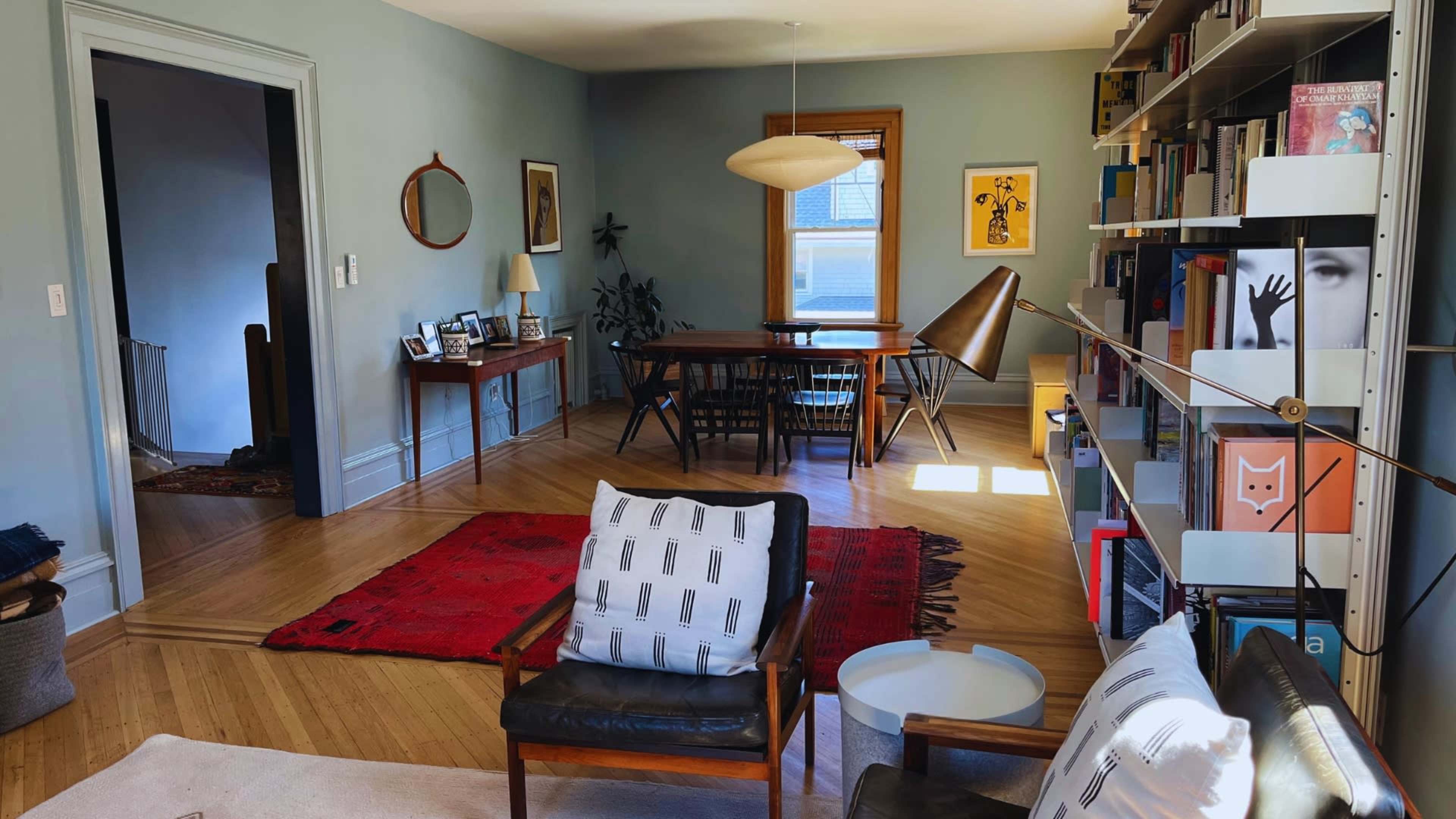 The image shows a living room with a dining area, featuring a wooden table, various chairs, a bookshelf, and a red area rug.
