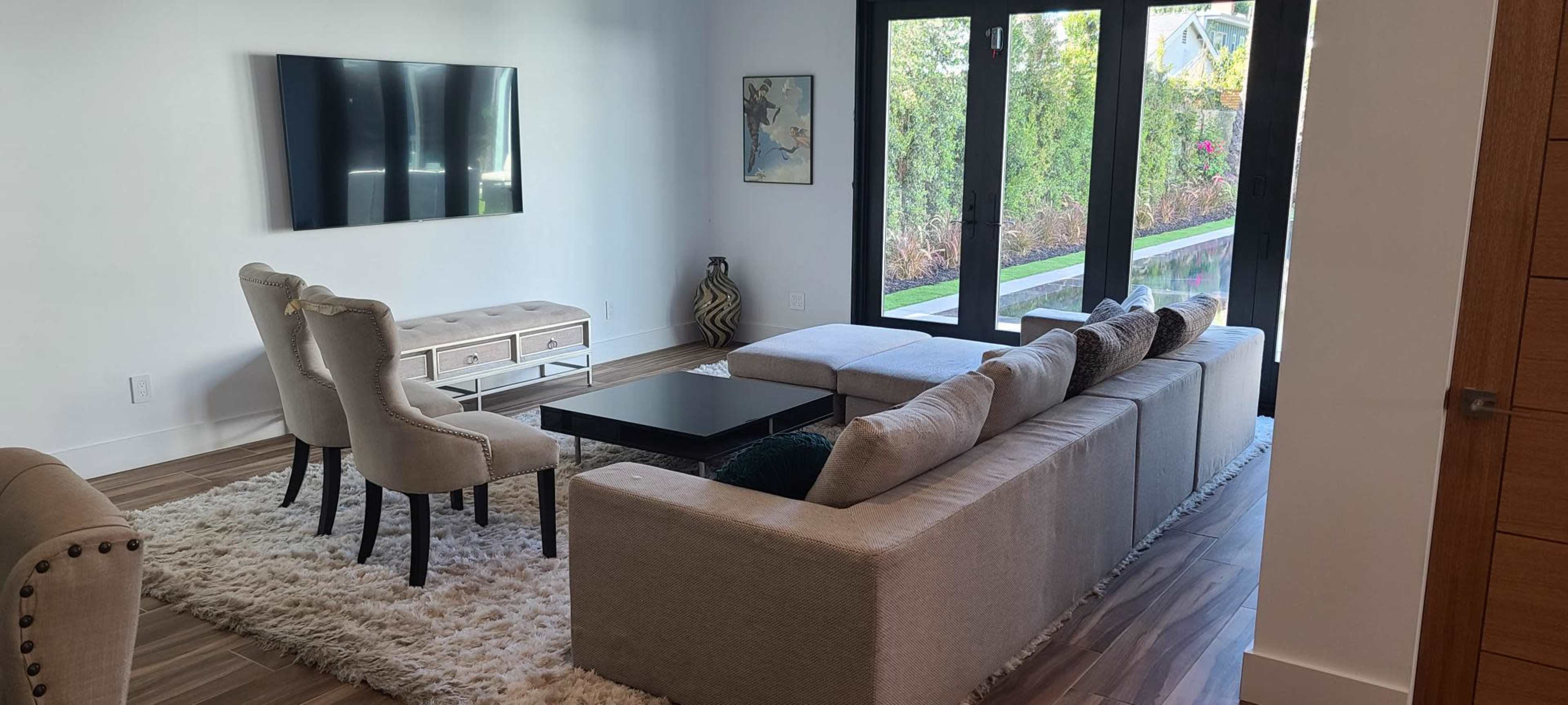A modern living room with a sectional sofa, coffee table, and a wall-mounted television, featuring large windows that provide a view of an outdoor space.