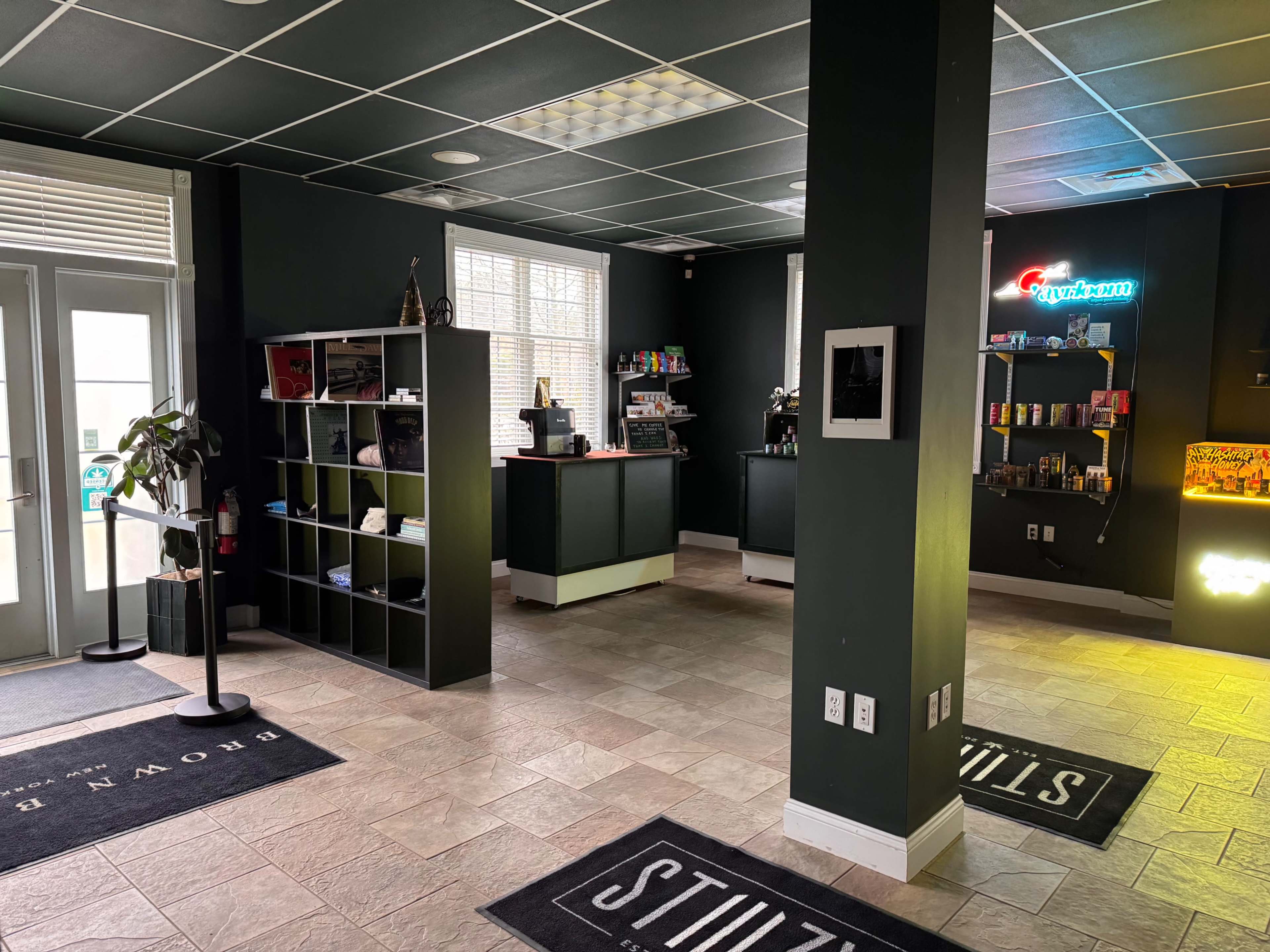 The image shows a modern interior space featuring dark walls, shelves displaying various items, and a reception area with a counter and several plants.