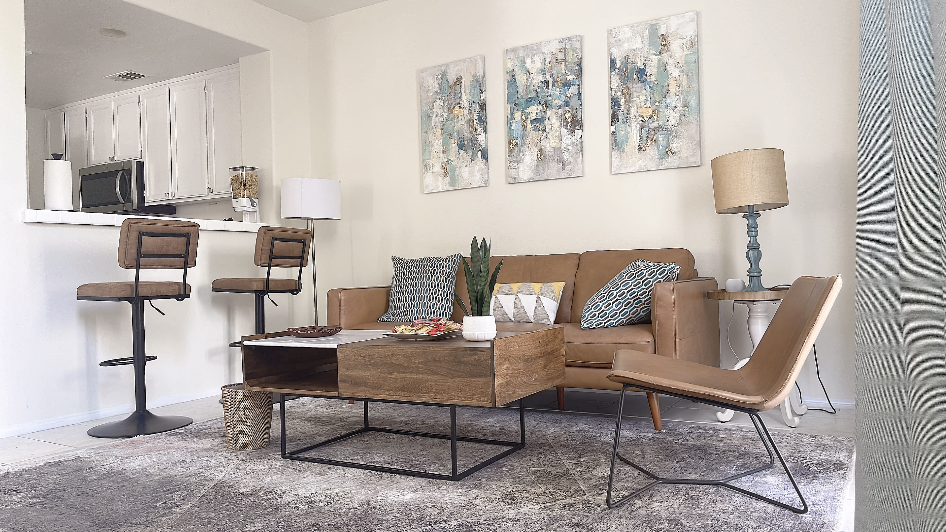 A modern living room with a brown leather sofa, two bar stools at a kitchen counter, and abstract art on the walls, alongside a coffee table and a potted plant.
