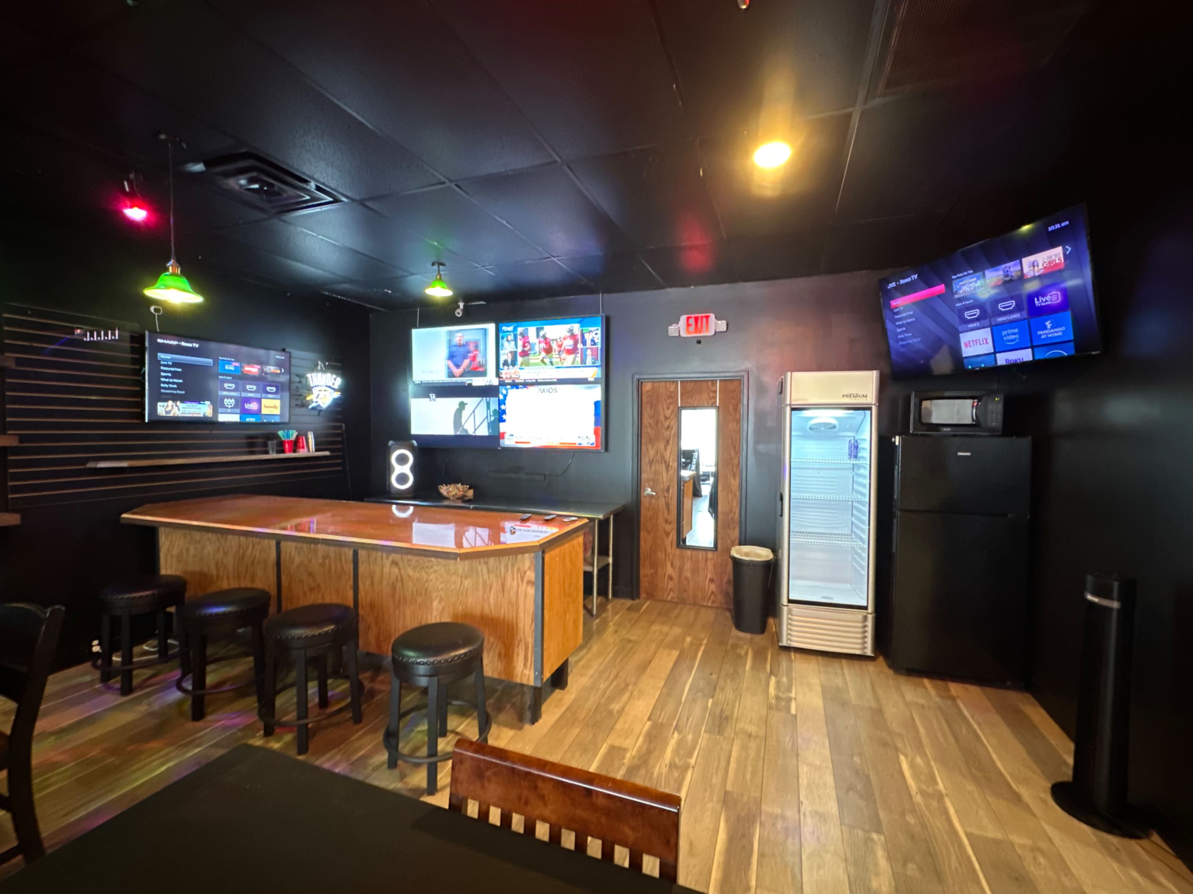 The image shows a modern bar area with multiple televisions on the walls, a wooden bar counter, and stools arranged in front.